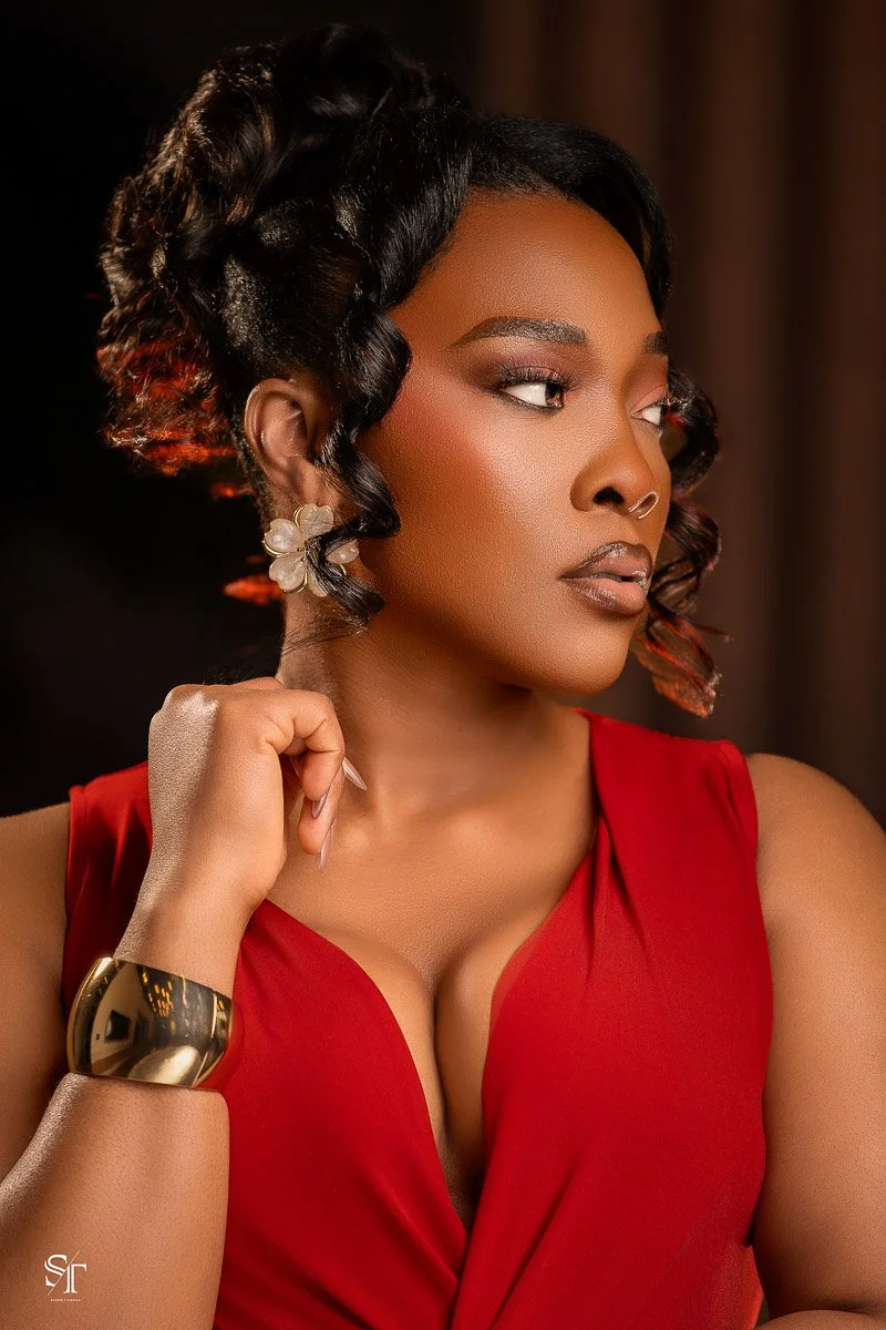 A woman with styled black hair and makeup, wearing a red sleeveless dress, a gold bracelet, floral earrings, and a nose ring, posing with her hand near her face against a dark background.