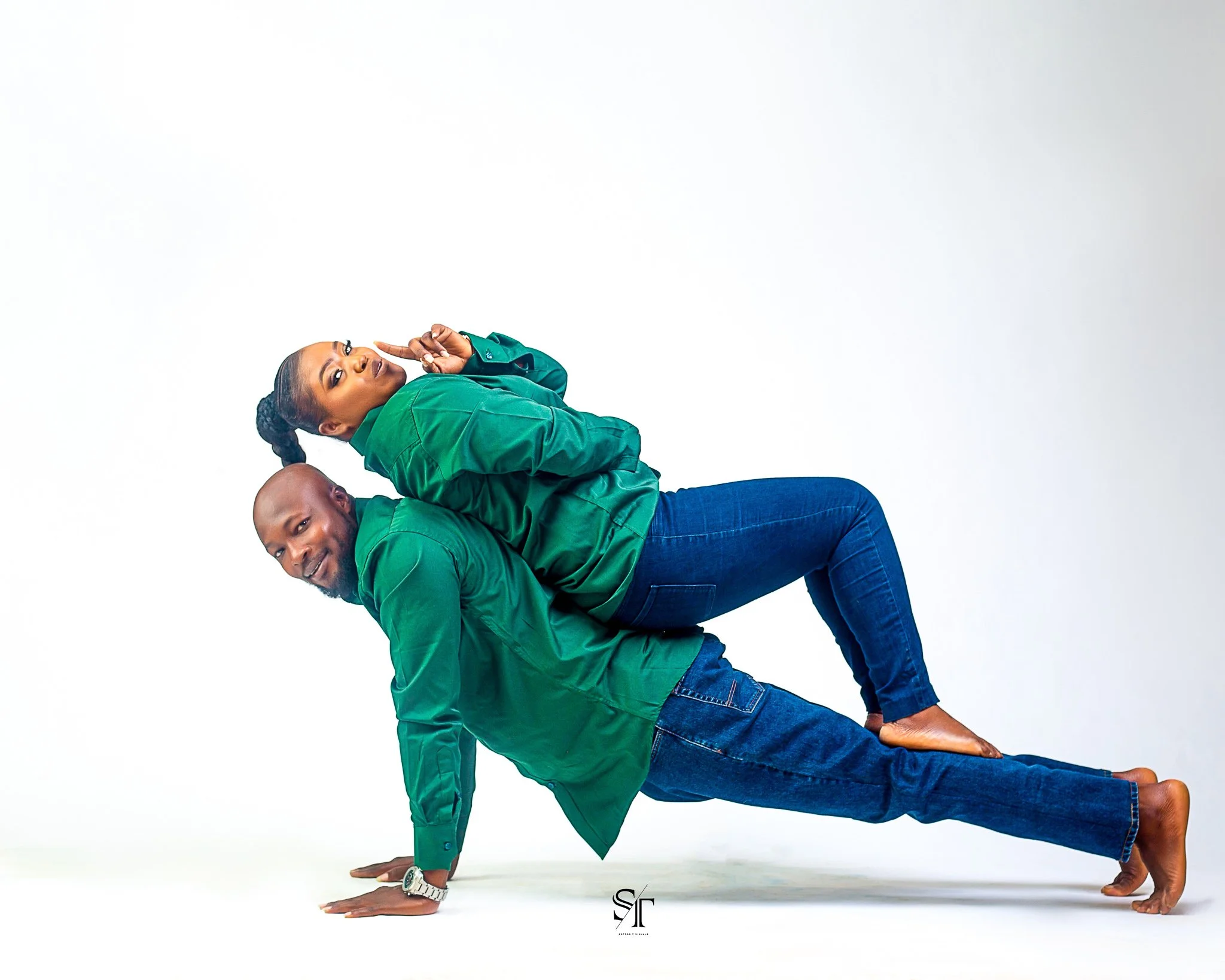 A man on hands and knees with a woman lying on his back, both smiling, wearing green shirts and blue jeans, against a plain white background.