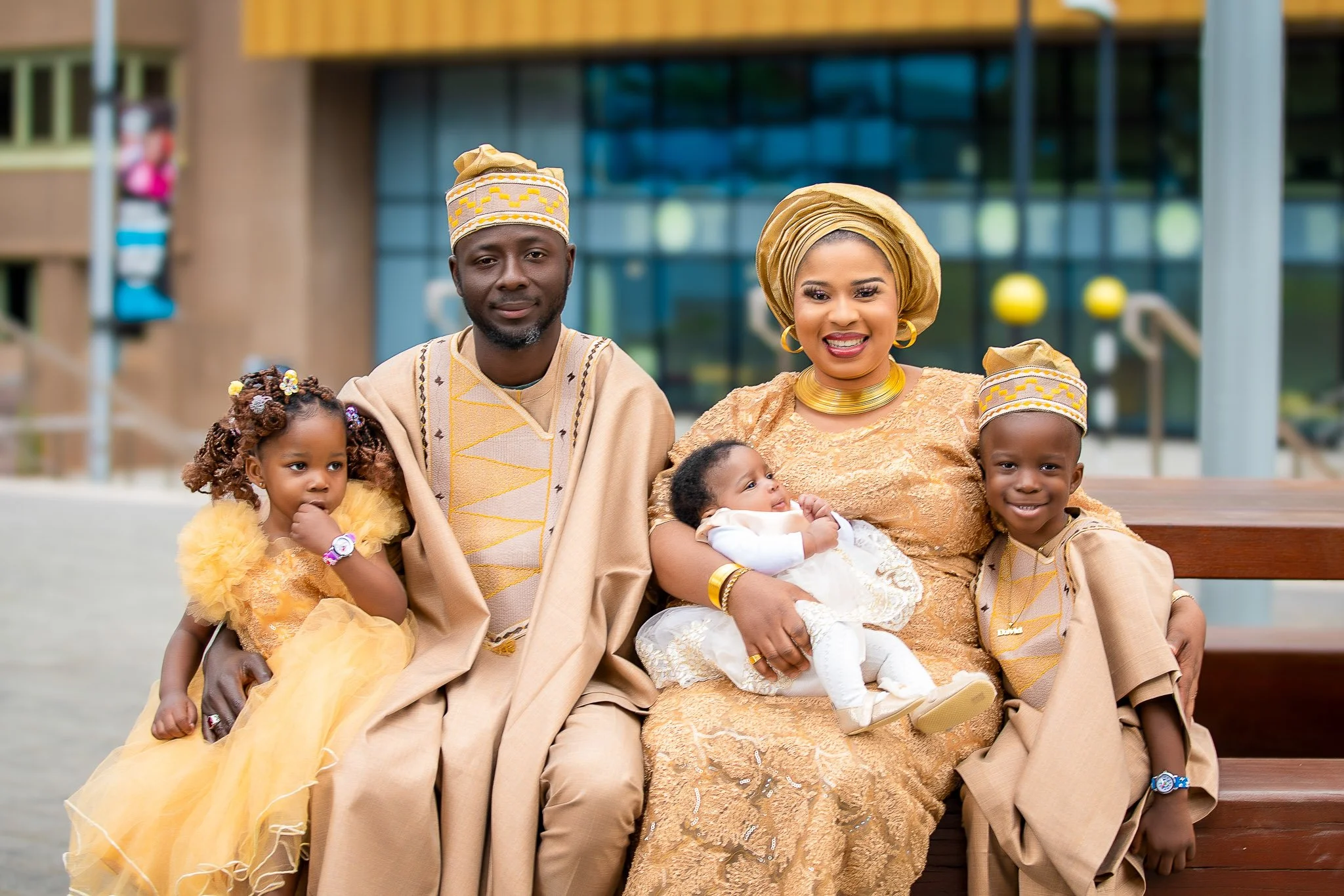A family of five dressed in traditional African attire sitting outdoors, with a modern building in the background.