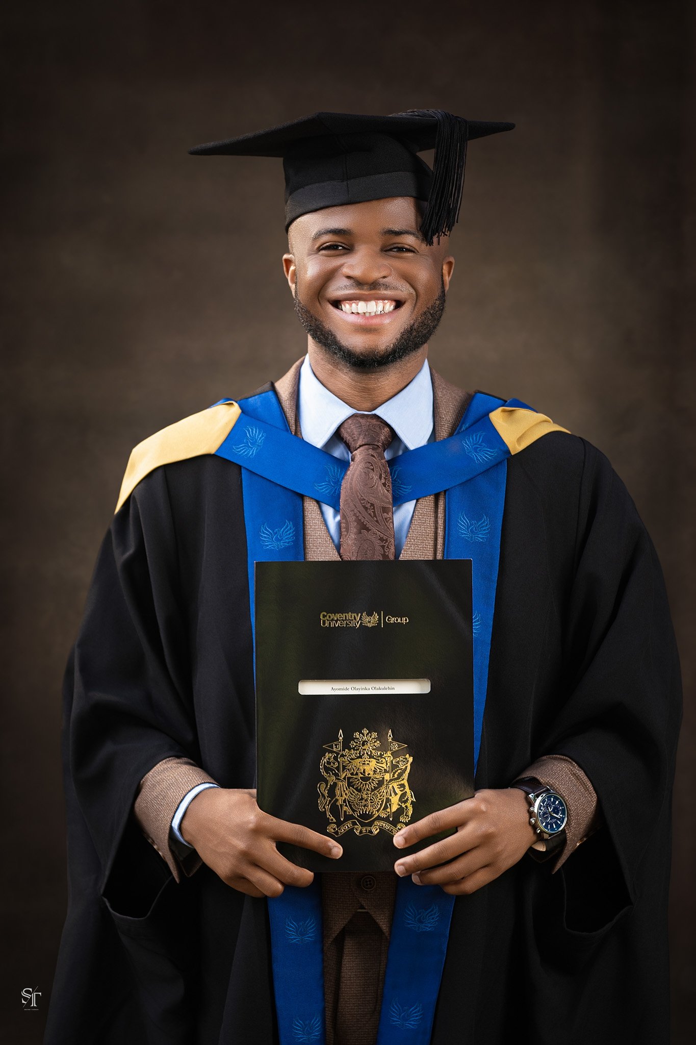 A smiling man in graduation robes and cap holding a diploma with a university crest, wearing a watch and a brown blazer.