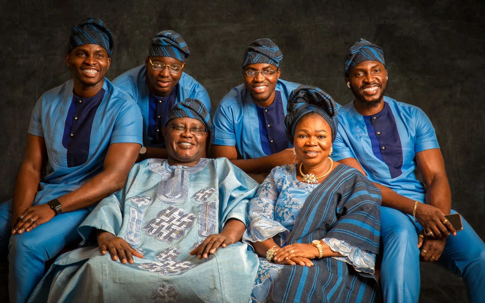 African family group dressed in traditional blue attire, sitting and standing against a dark background, smiling for the camera.