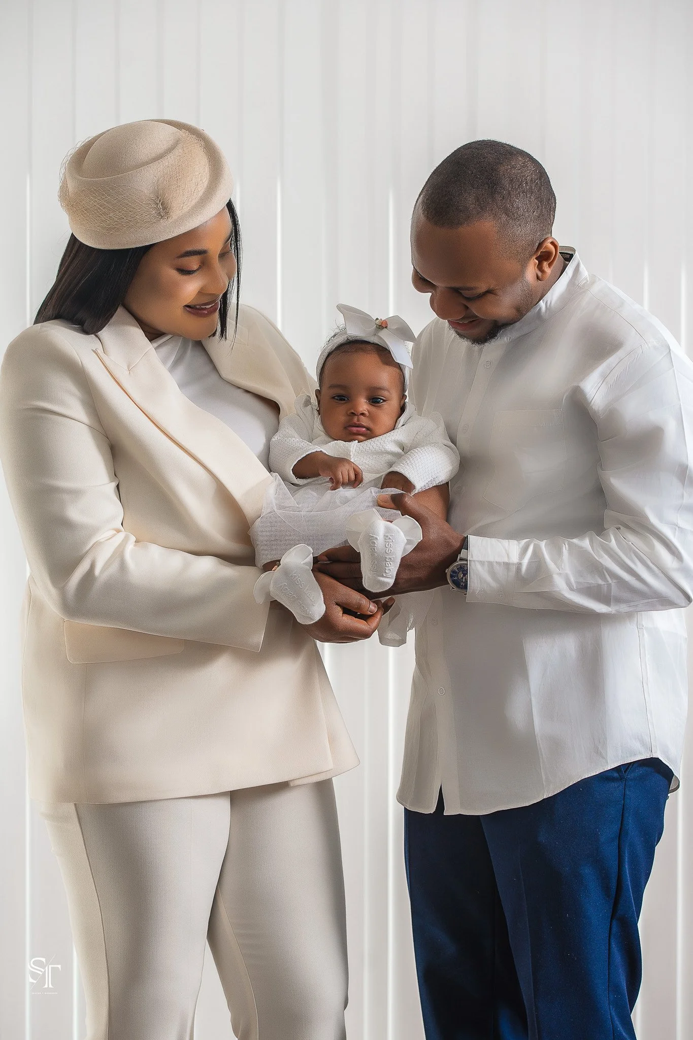 A family holding a baby girl dressed in white, with a woman in a beige suit and hat, and a man in a white shirt and blue pants, standing against a white paneled background.