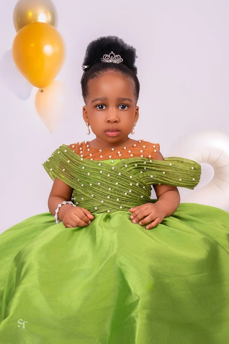 A young girl dressed in a green and orange princess gown with pearl embellishments, wearing jewelry and a tiara, sitting in front of a white background with gold and white balloons.