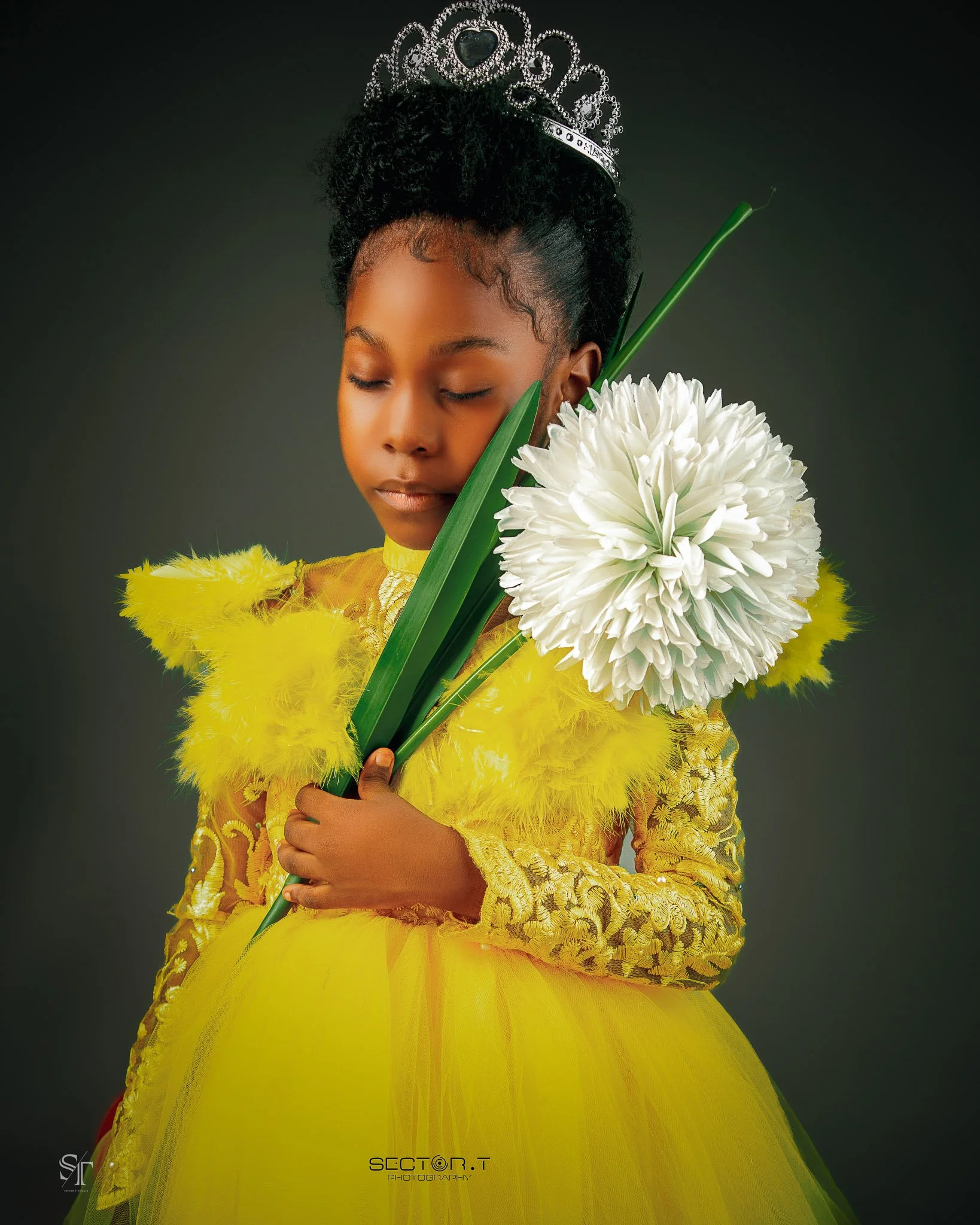 Young girl in a yellow dress with lace details, wearing a tiara, holding a white flower and green leaves, with a serene expression and eyes closed.