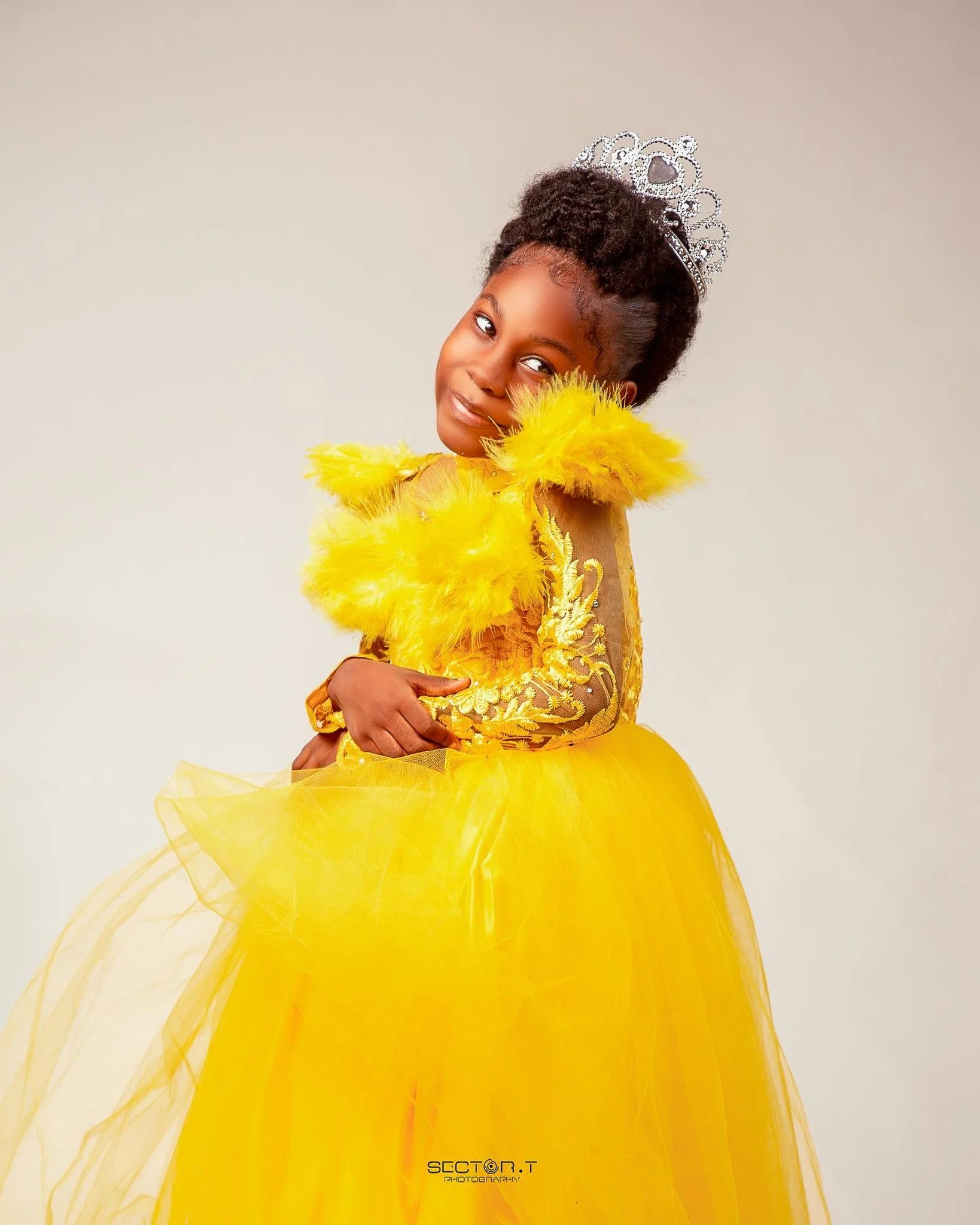 A young girl wearing a yellow princess dress with feather and floral details, a yellow fluffy boa, and a silver tiara, posing against a light neutral background.