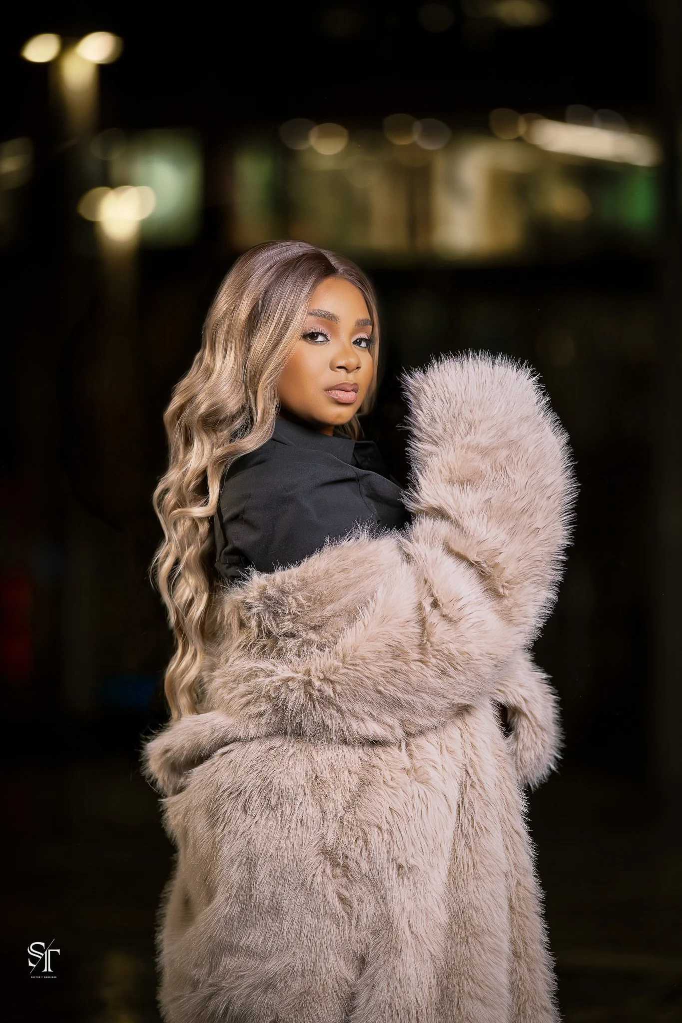 A woman with long, wavy blonde hair wearing a black top and a beige faux fur coat, standing against a blurred night city background.