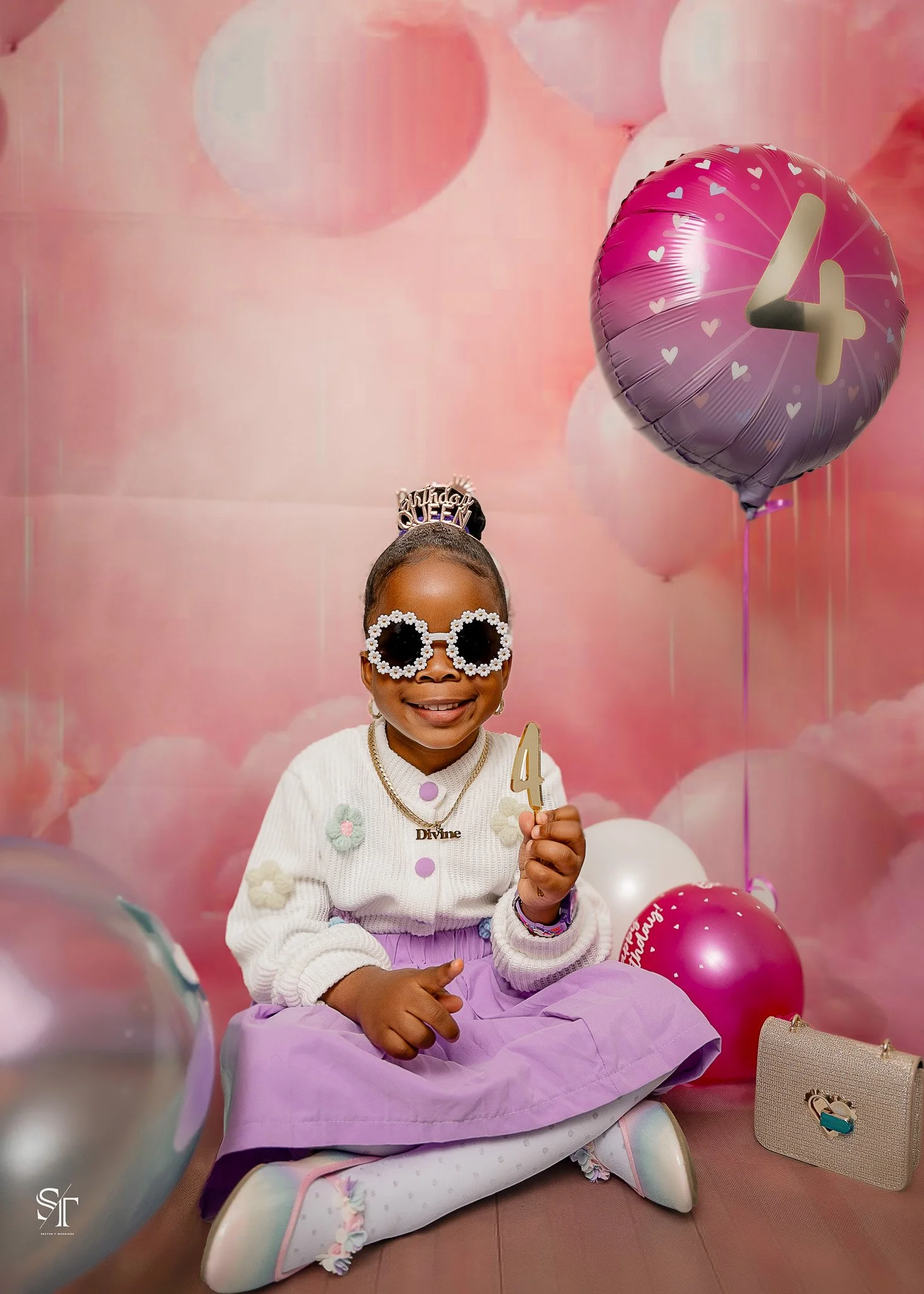 Young girl sitting on a pastel-colored cushion, wearing large sunglasses, a crown that says 'Birthday Queen,' holding a gold '4' candle, with balloons in pink, purple, and white background and floor, and a small beige purse nearby.