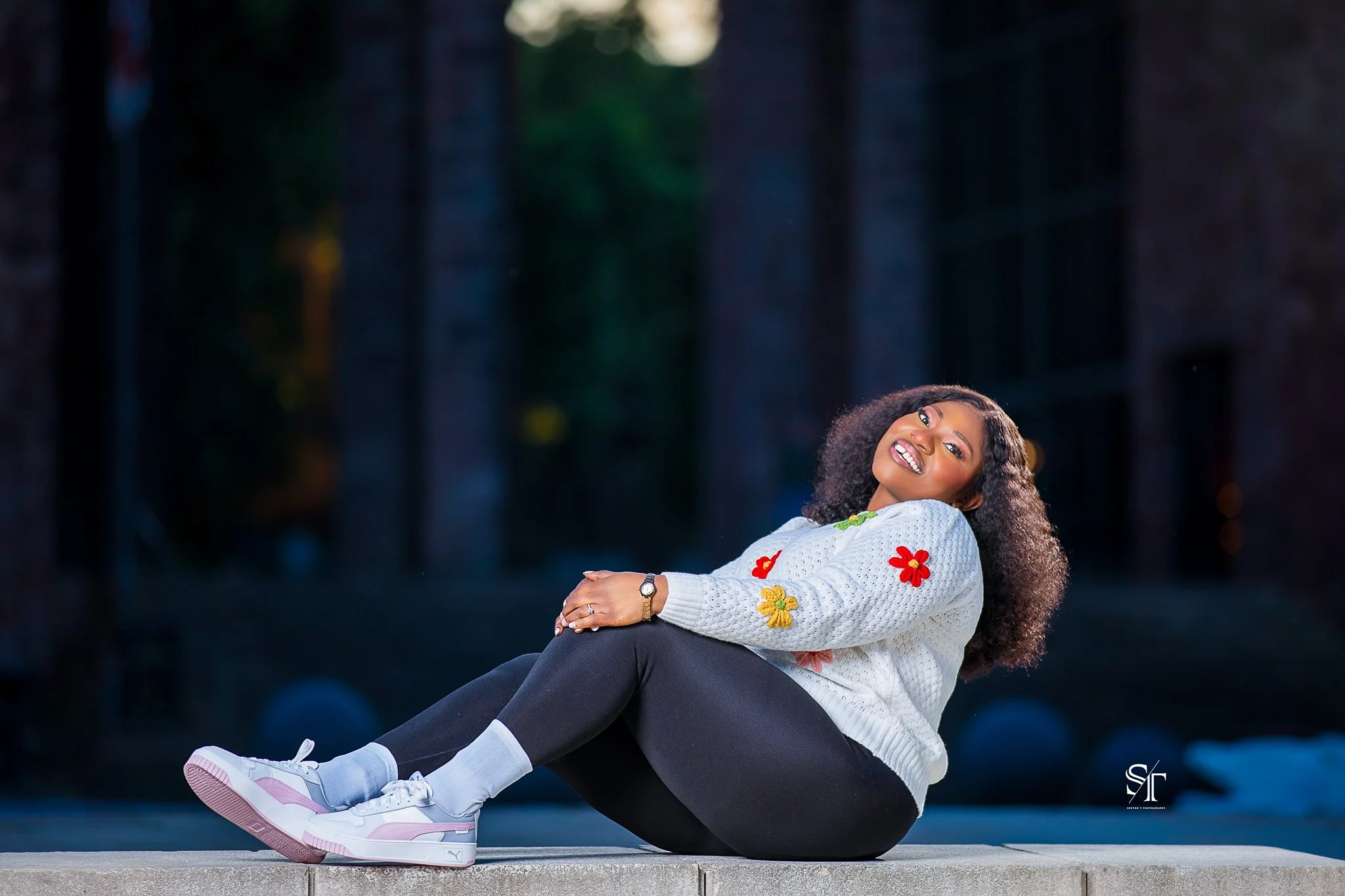 Young woman sitting on a ledge outdoors at night, smiling with curly hair, wearing a white sweater with colorful flower patches, black leggings, pink and white sneakers, and a watch.