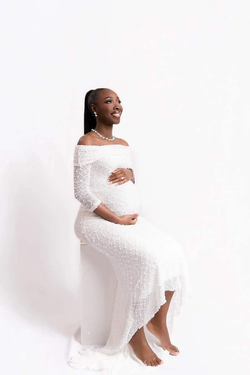 Pregnant woman in a white off-shoulder dress sitting against a plain white background, smiling and holding her belly.