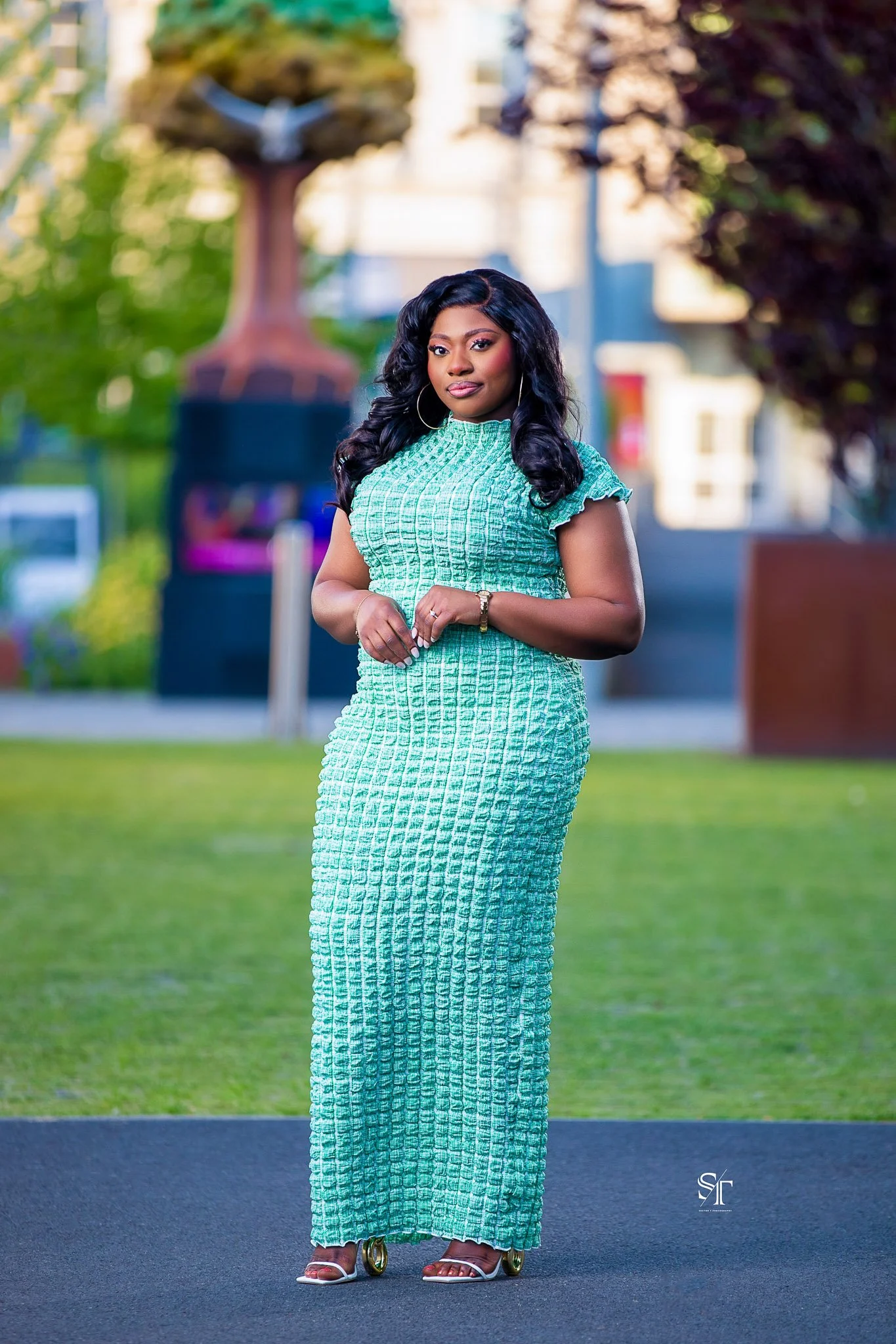A woman standing outdoors on a grassy area with trees and a sculpture in the background, wearing a green textured dress and gold high heels.
