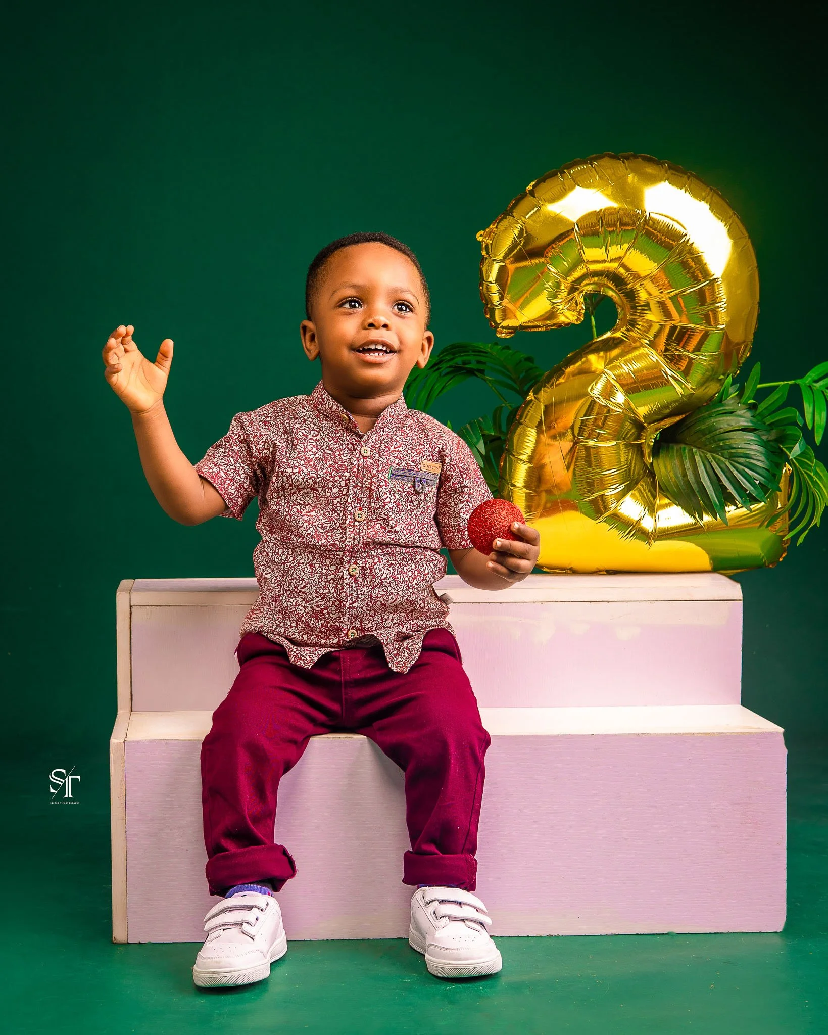 A young boy sitting on a pink bench, holding a red ball, waving with his right hand, celebrating his second birthday, with gold balloon shaped as the number 2 and green leaves in the background.
