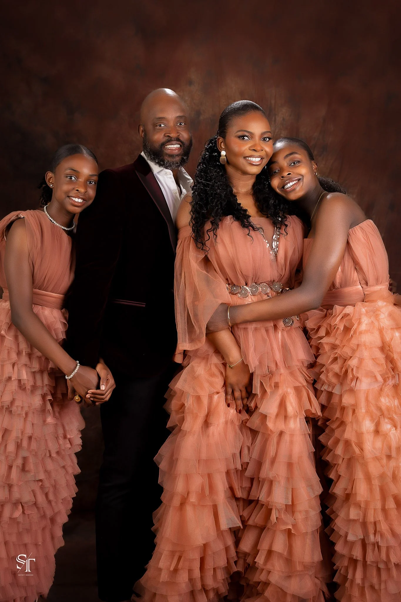 A family portrait featuring two adult women, a man, and two young girls, all dressed in matching peach-colored ruffled dresses and formal attire, posing together against a dark brown studio backdrop.