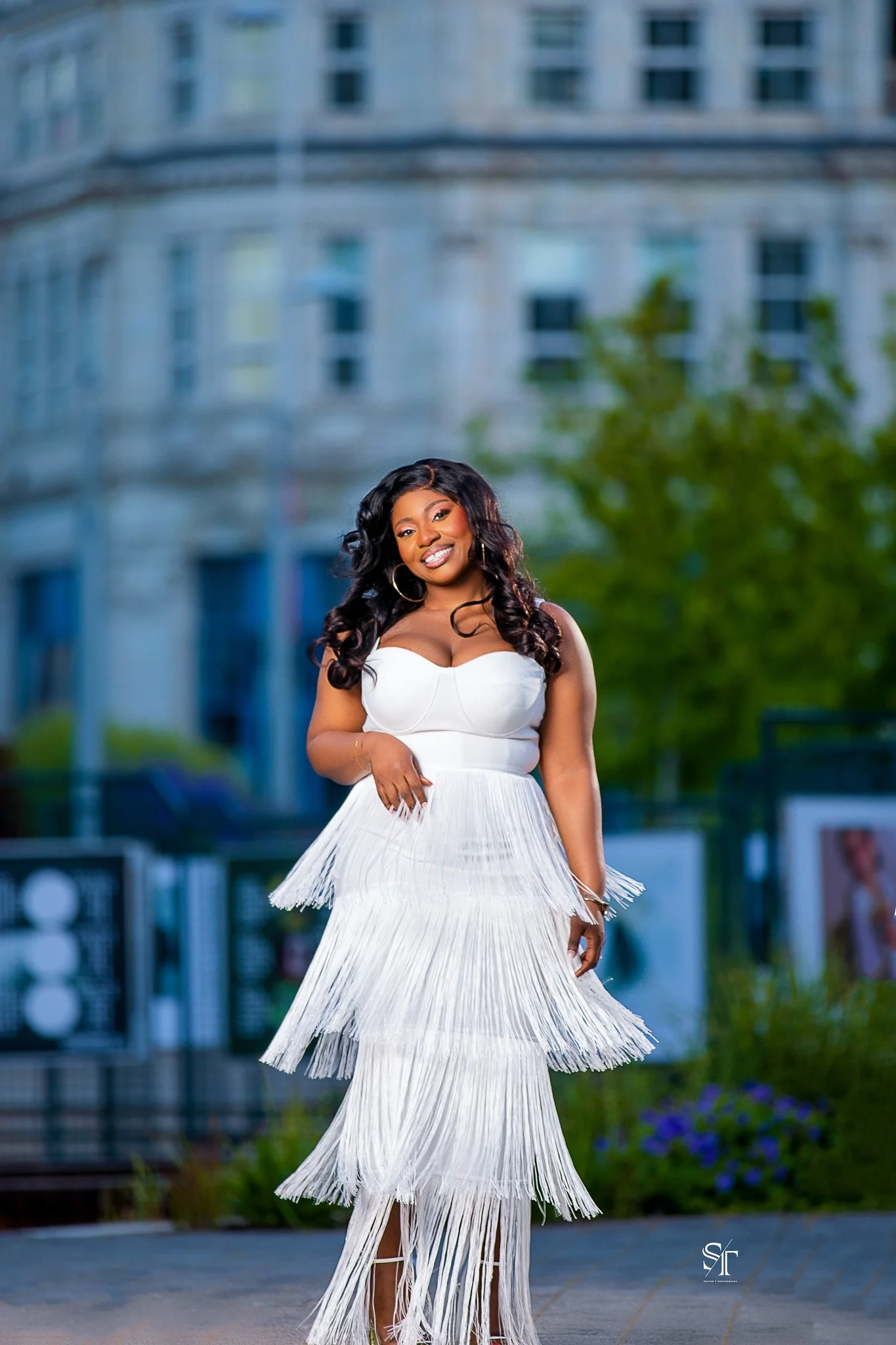 Young woman in a white, strapless, tiered fringe dress smiling outdoors in front of a city building with greenery.