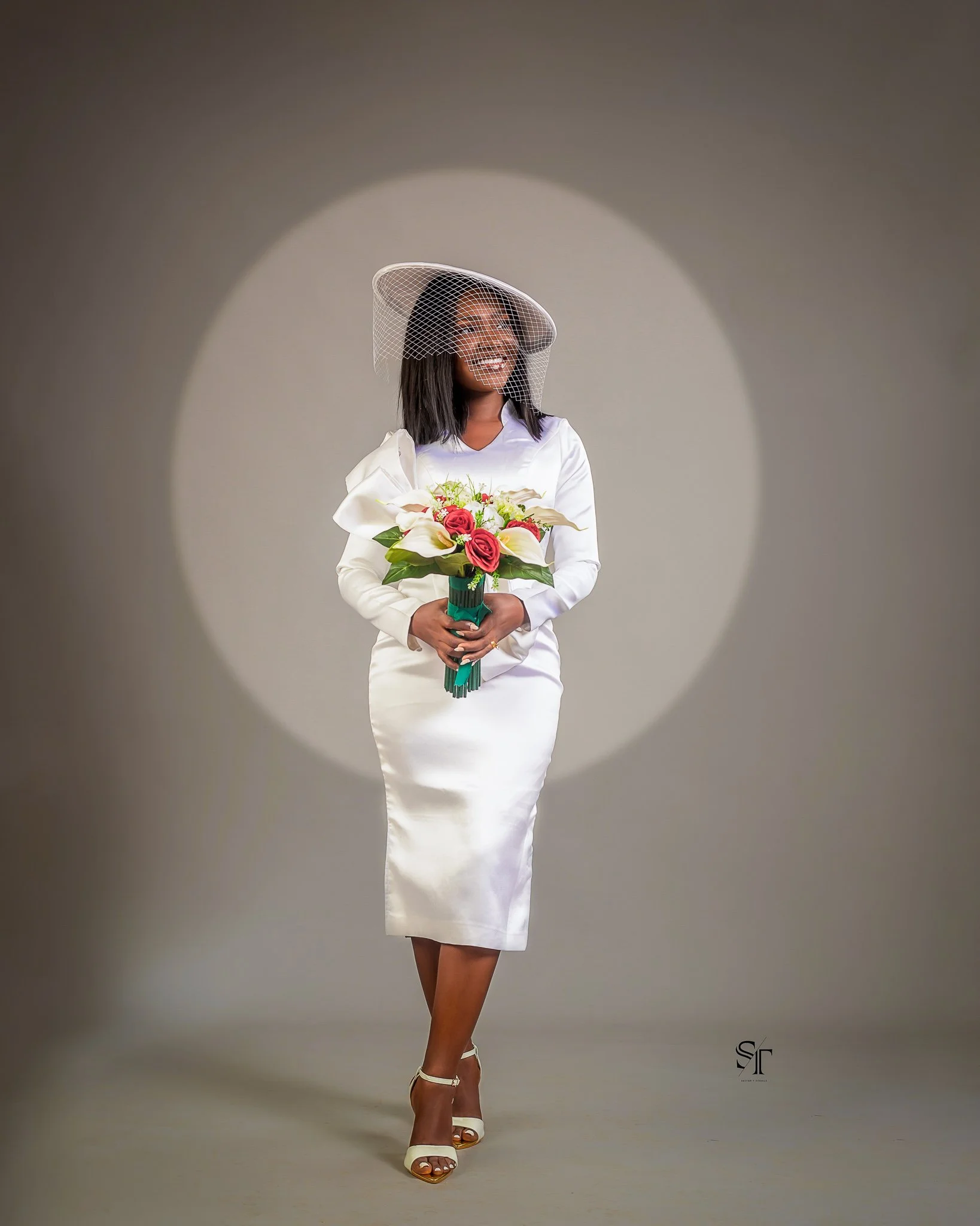 A woman in a white dress with a matching wide-brimmed hat decorated with a veil, holding a colorful bouquet of flowers, standing on a plain background with a spotlight effect.