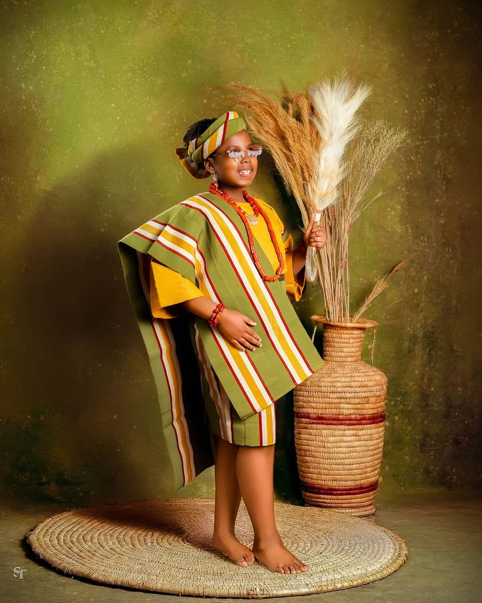 A young girl dressed in traditional African clothing, standing barefoot on a woven rug, holding a bundle of dried pampas grass in front of a green background, with a large woven vase behind her.