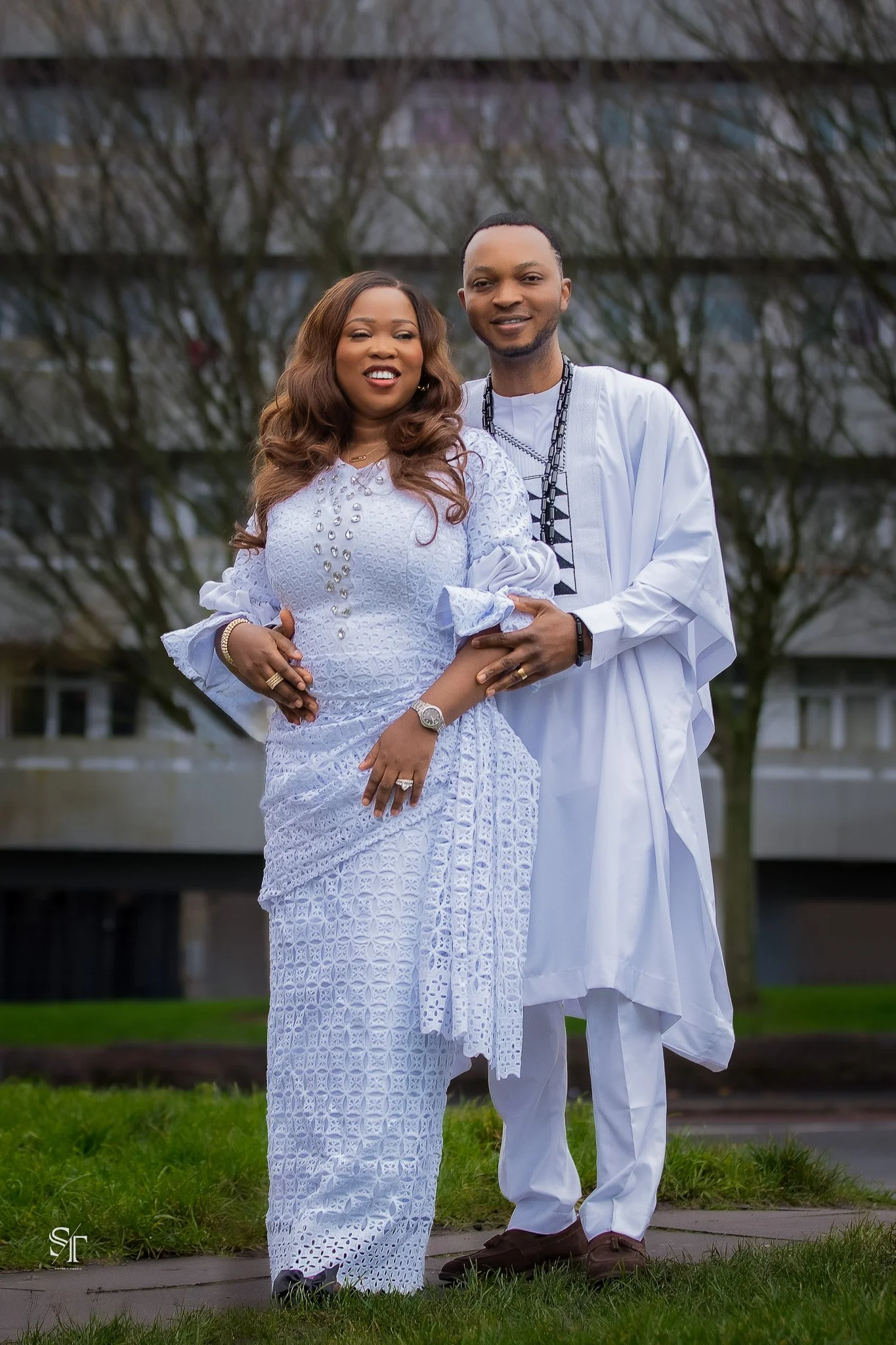 A couple dressed in traditional African attire standing outside on grass with trees and a building in the background.