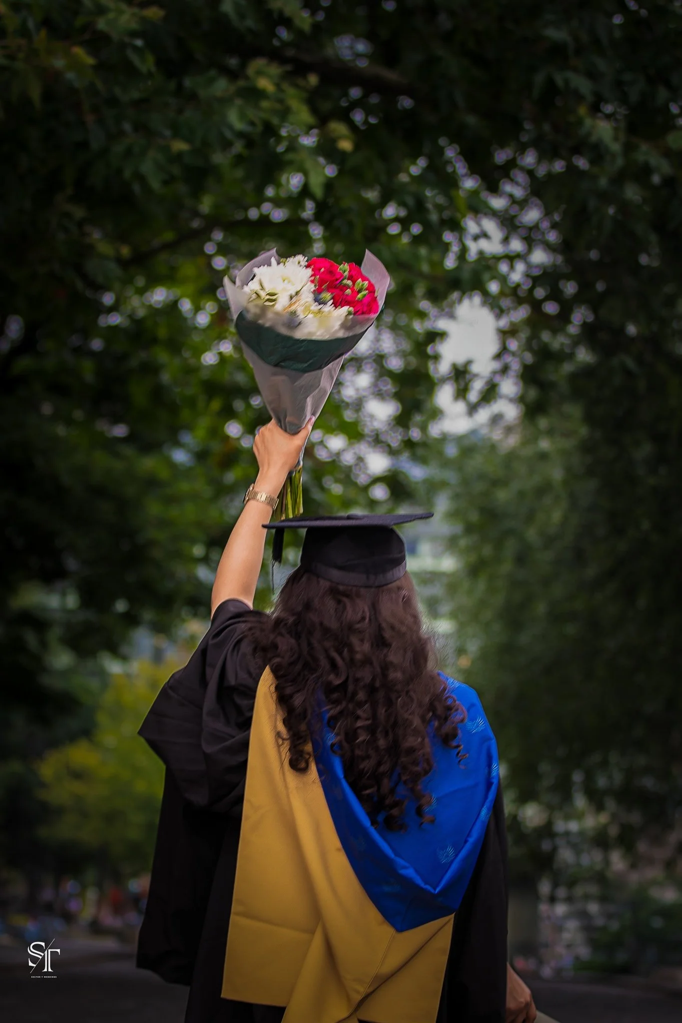 Graduate in cap and gown holding bouquet of flowers outdoors.
