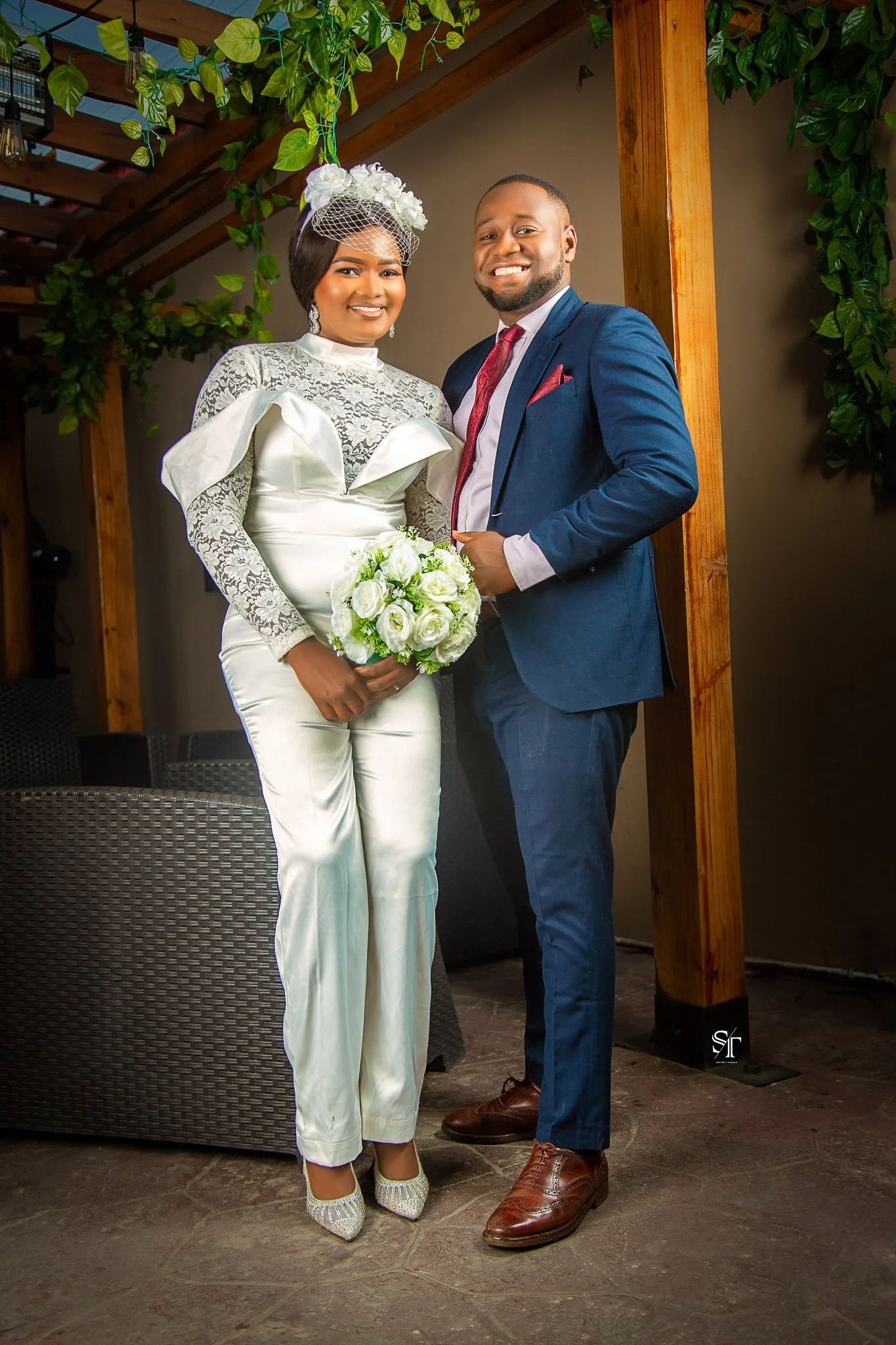 A bride in a white satin and lace wedding gown holding a bouquet of white roses, standing next to a groom in a navy blue suit, white shirt, and red tie at a wedding venue decorated with green plants and wooden beams.