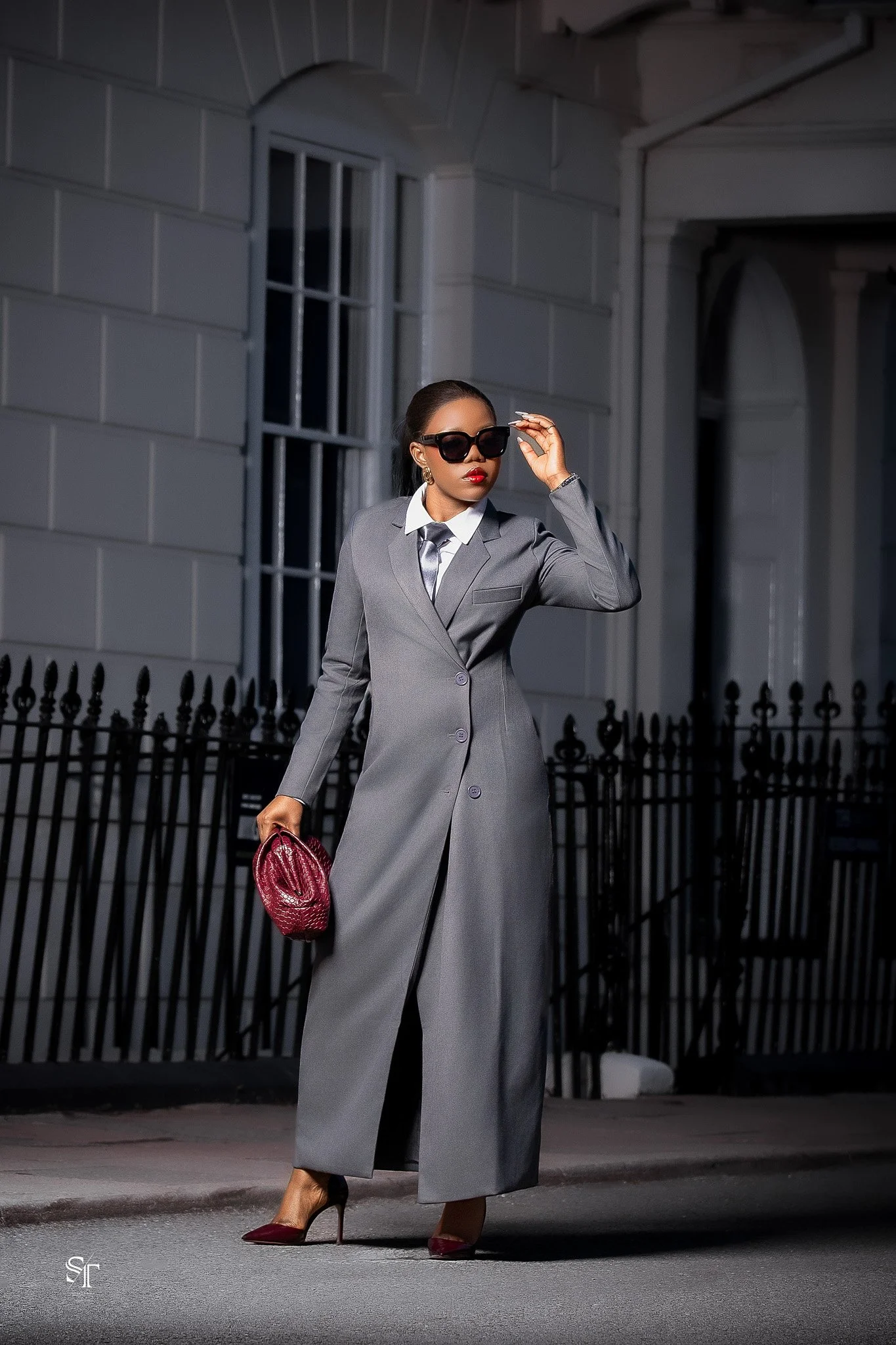 Woman in gray suit, black sunglasses, red high heels, standing outside near white building with barred window, holding a red handbag.