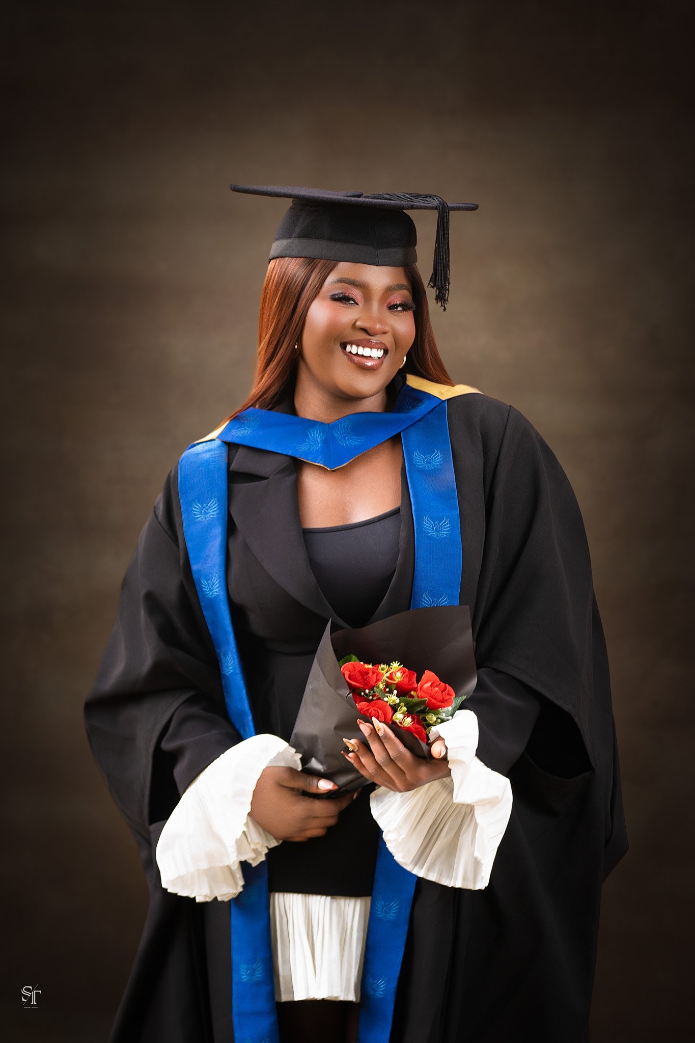A woman in a graduation cap and gown holding a bouquet of red and yellow flowers, smiling at the camera.