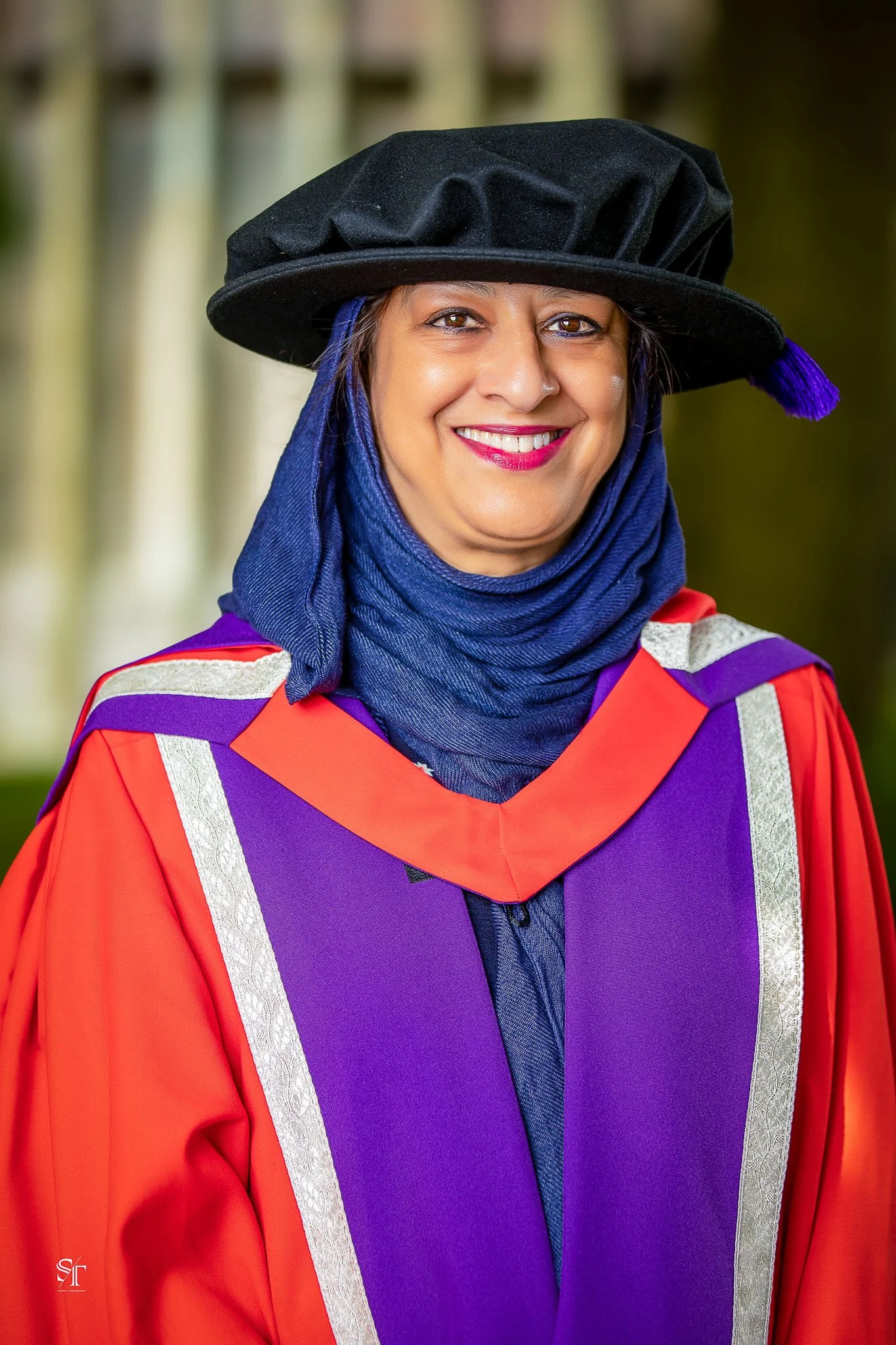A woman in academic regalia, wearing a black academic cap with purple tassel, purple and red gown with silver accents, and a blue scarf, smiling outdoors in front of a blurred green background.