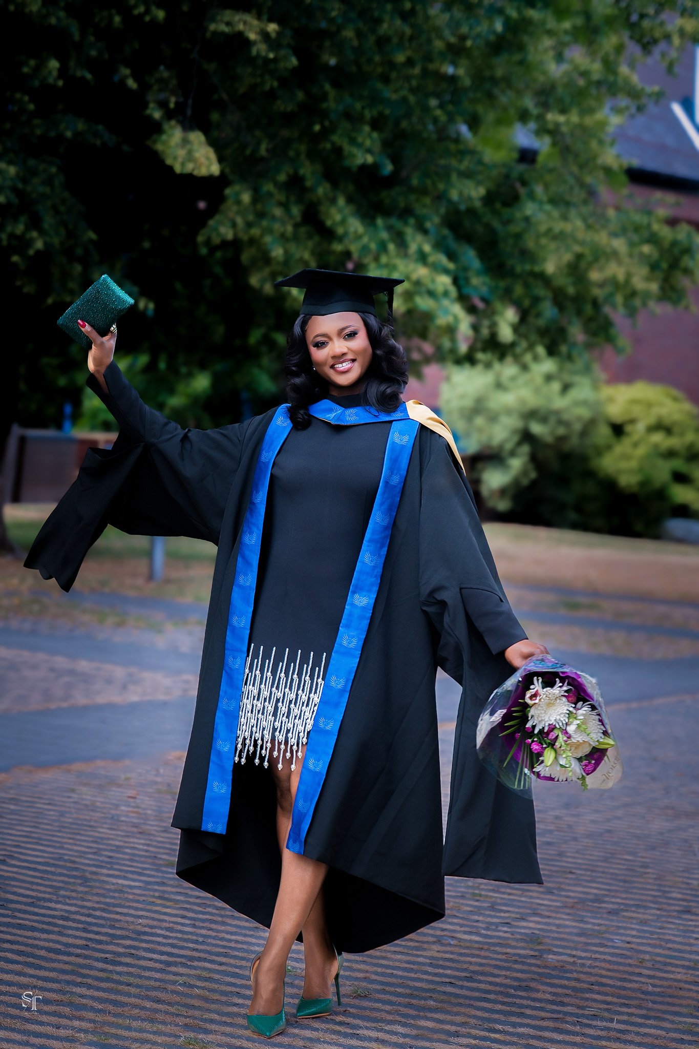 A woman in graduation cap and gown holding a bouquet of flowers and a clutch purse, standing outdoors with trees in the background.