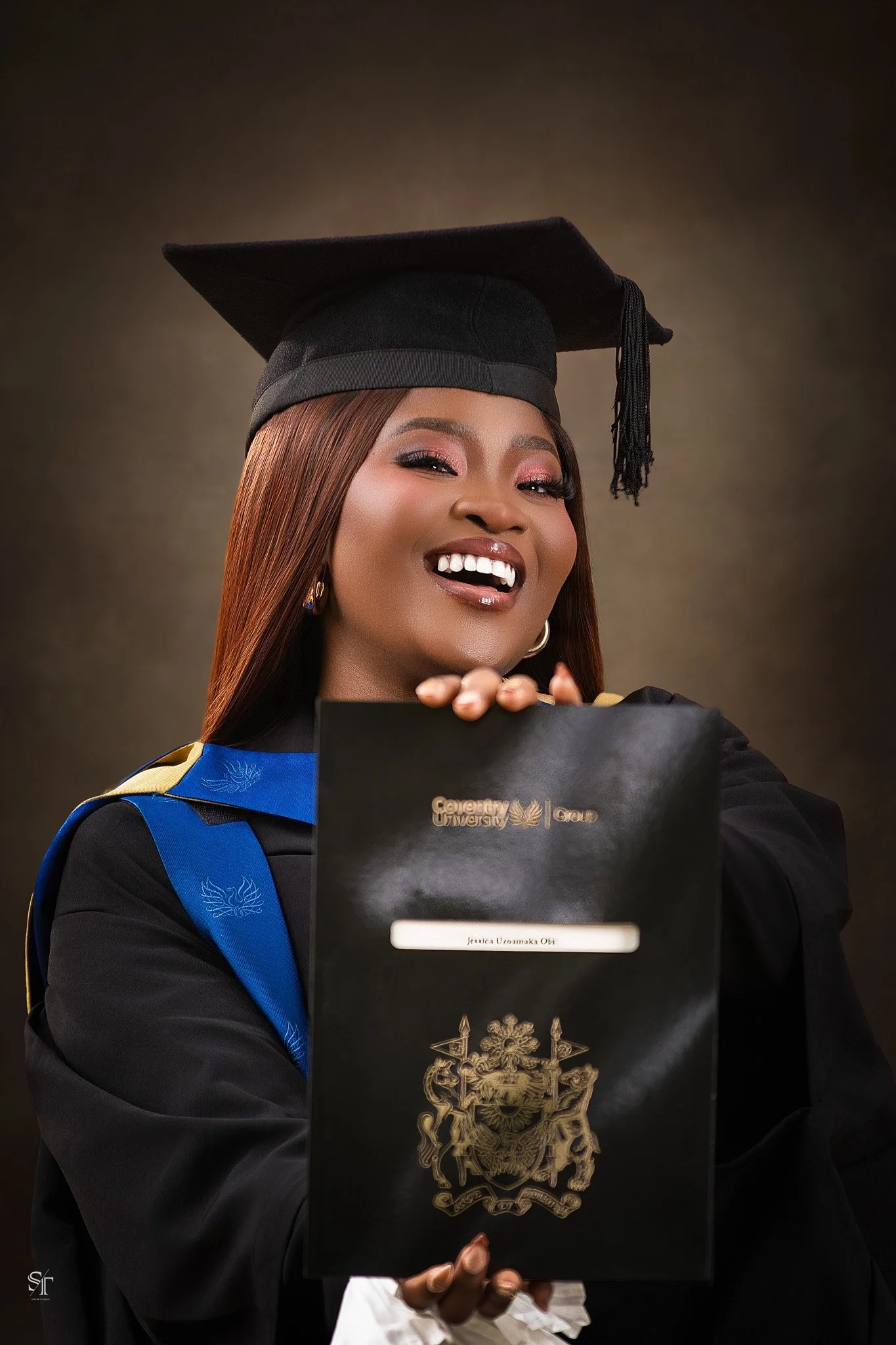 Smiling woman in graduation gown and cap holding her diploma in front of a dark background