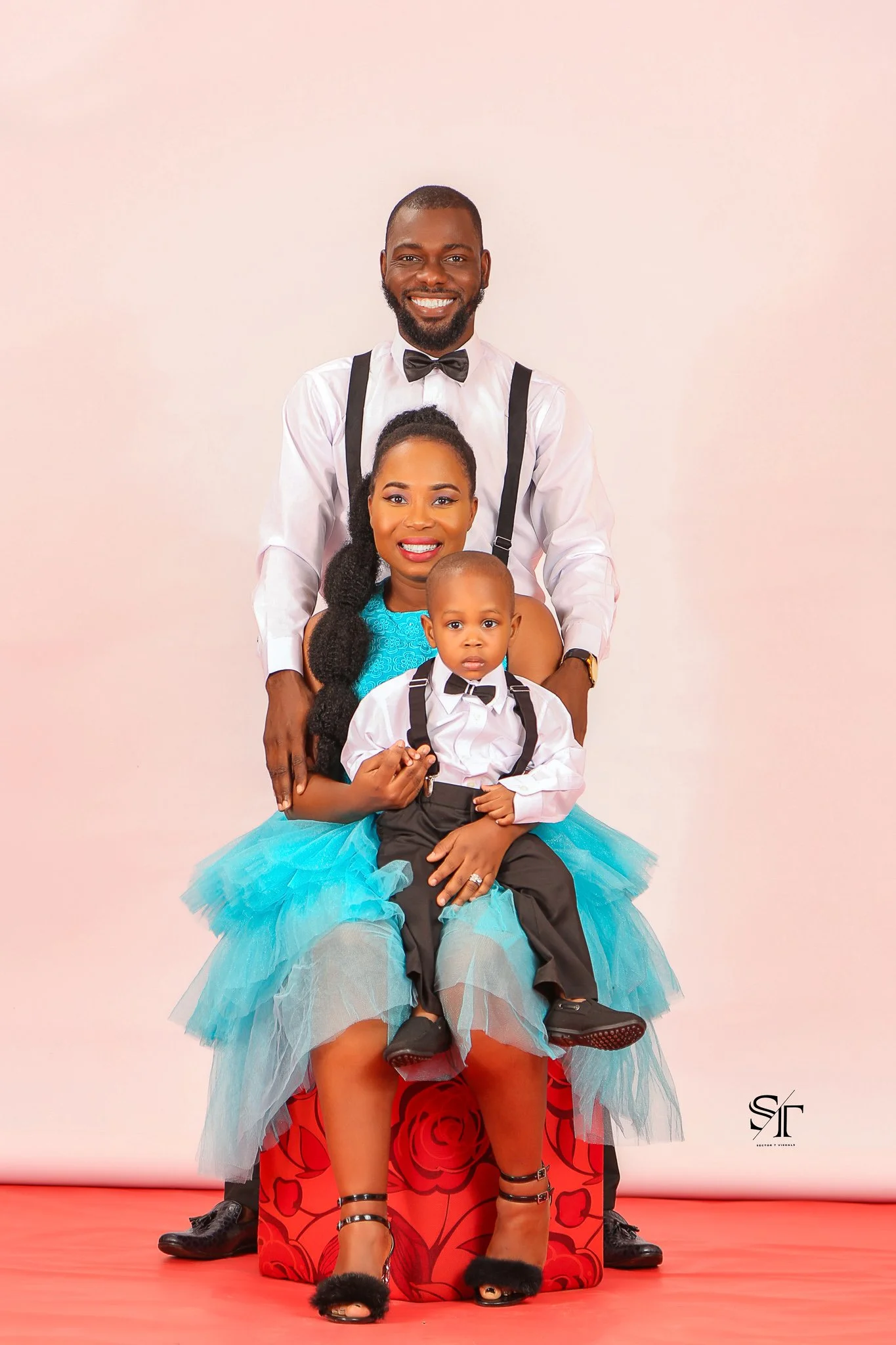 A family portrait featuring a man, woman, and a young boy dressed in formal attire, with the woman seated on a red floral stool and the man standing behind her, set against a plain light-colored background.