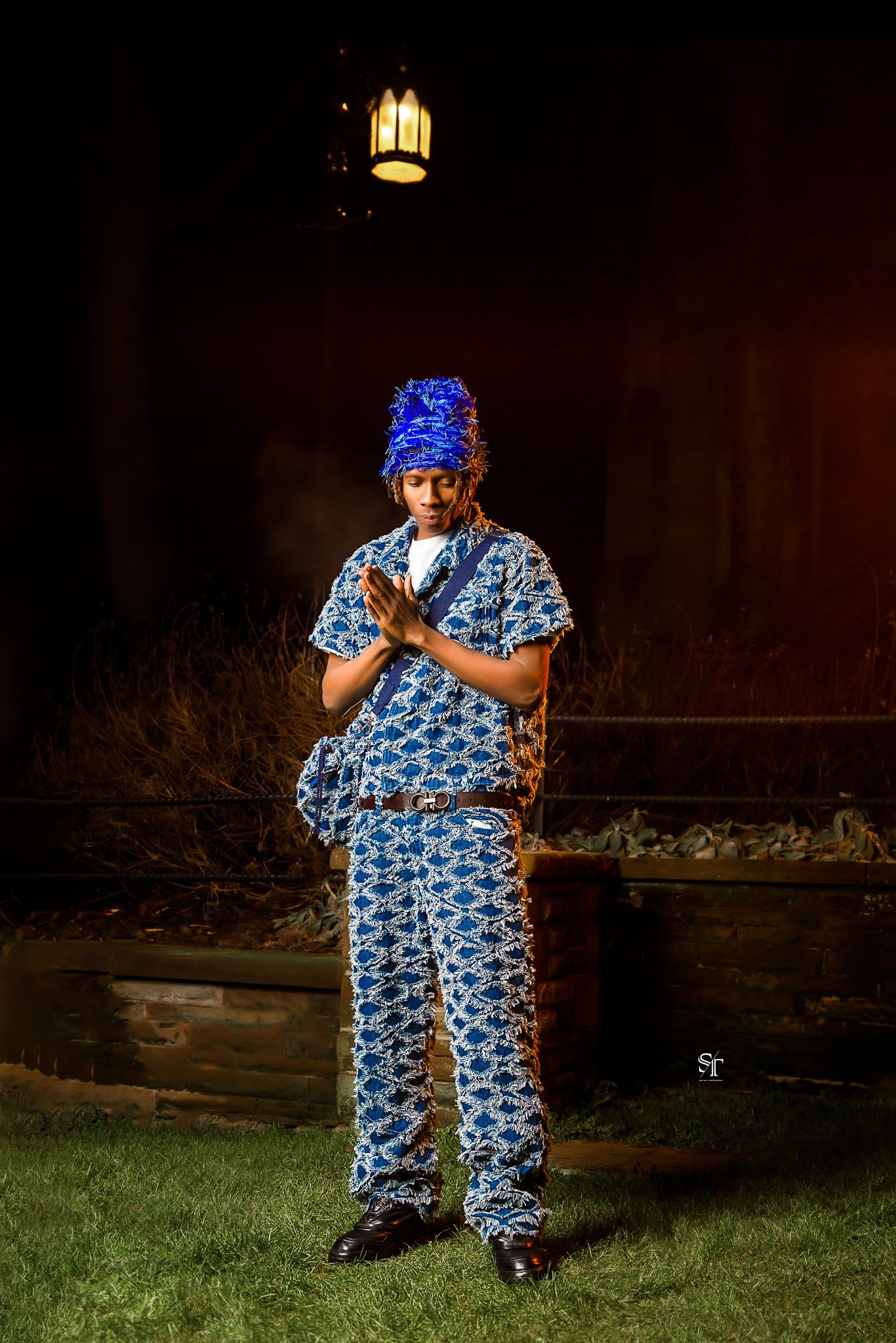 A person dressed in a blue and white patterned outfit, wearing a matching blue feathered headpiece, standing outdoors at night with hands together in a praying or meditative pose.