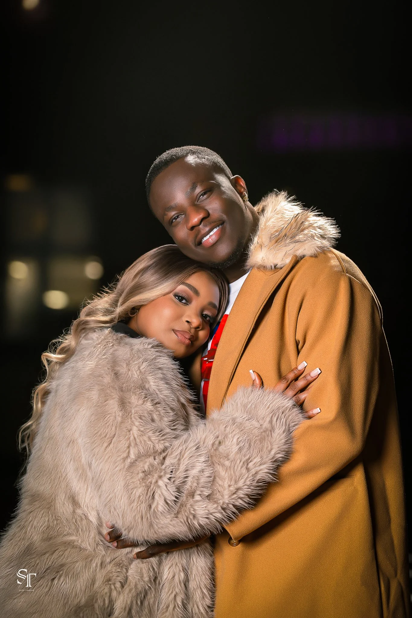 A young couple embracing at night, wearing warm coats with fur trim, smiling and looking at the camera.