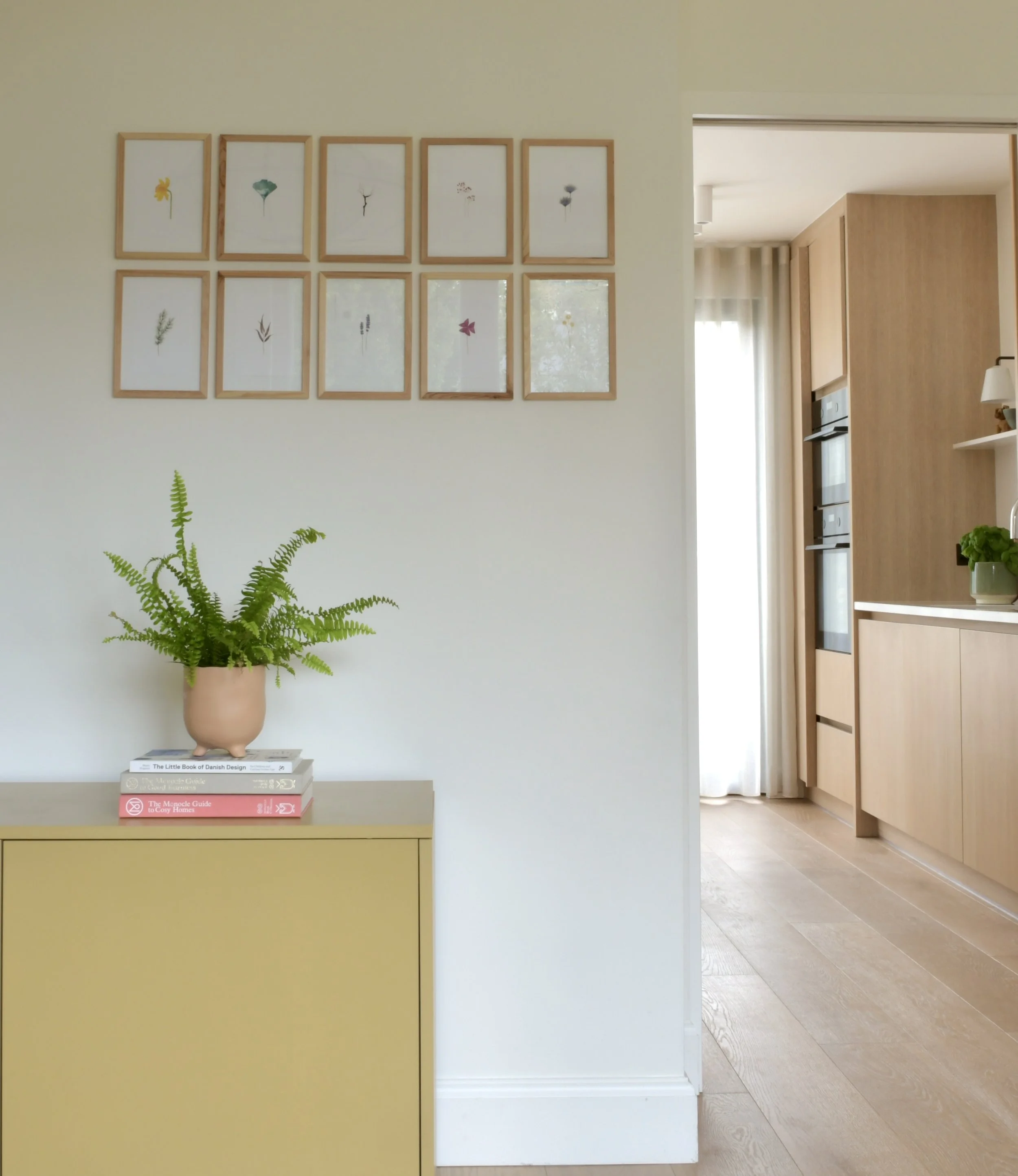 A living room with a yellow sideboard, a stack of books on top, a potted fern plant in a pink planter, white walls, and twelve framed botanical prints arranged in three rows and four columns. An open doorway reveals a kitchen area with wooden cabinet