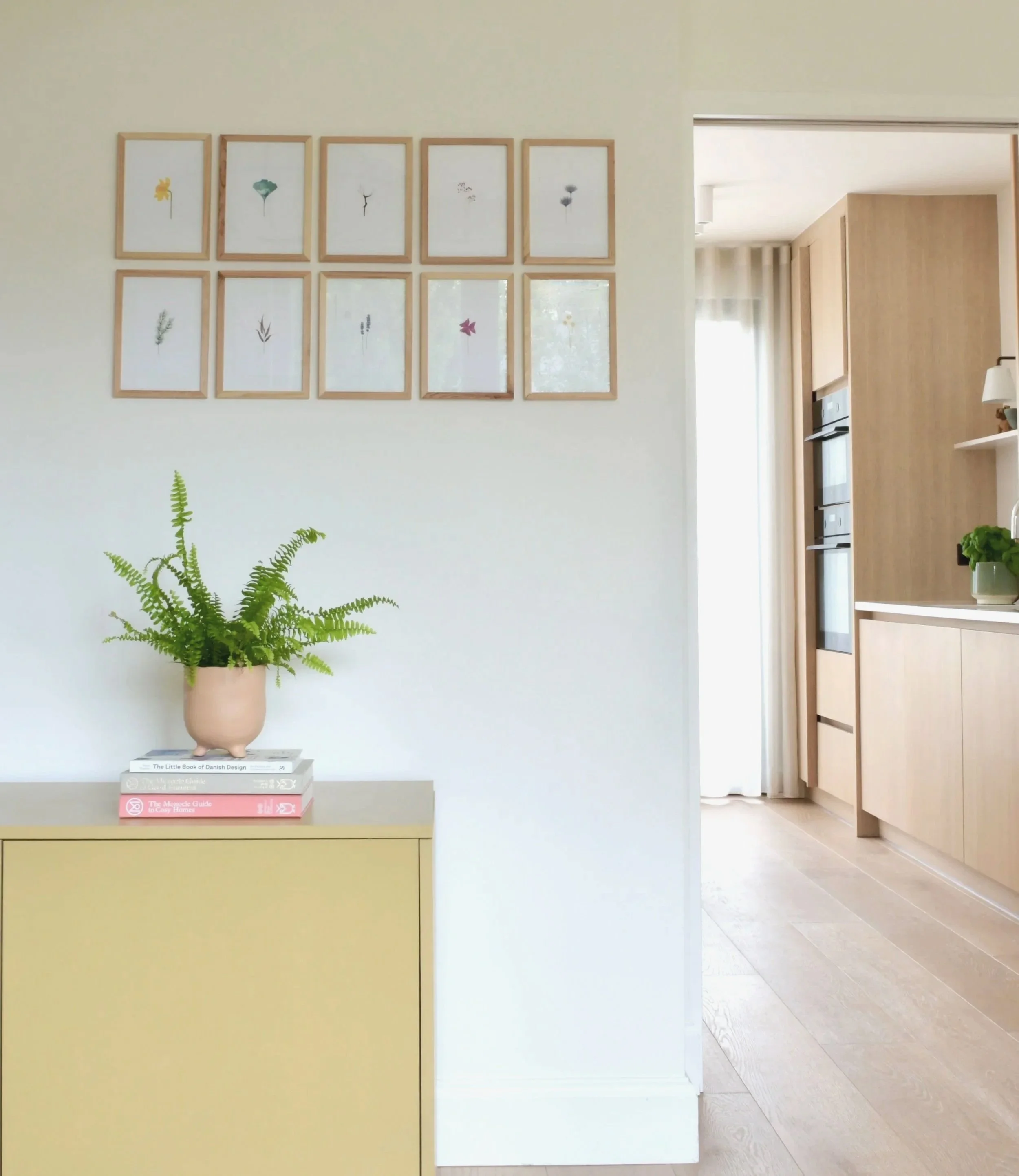 Interior view of a modern home featuring a wall display of framed botanical prints, a yellow side table with books and a potted fern plant, and a doorway leading to a kitchen area with wooden cabinets and natural lighting.