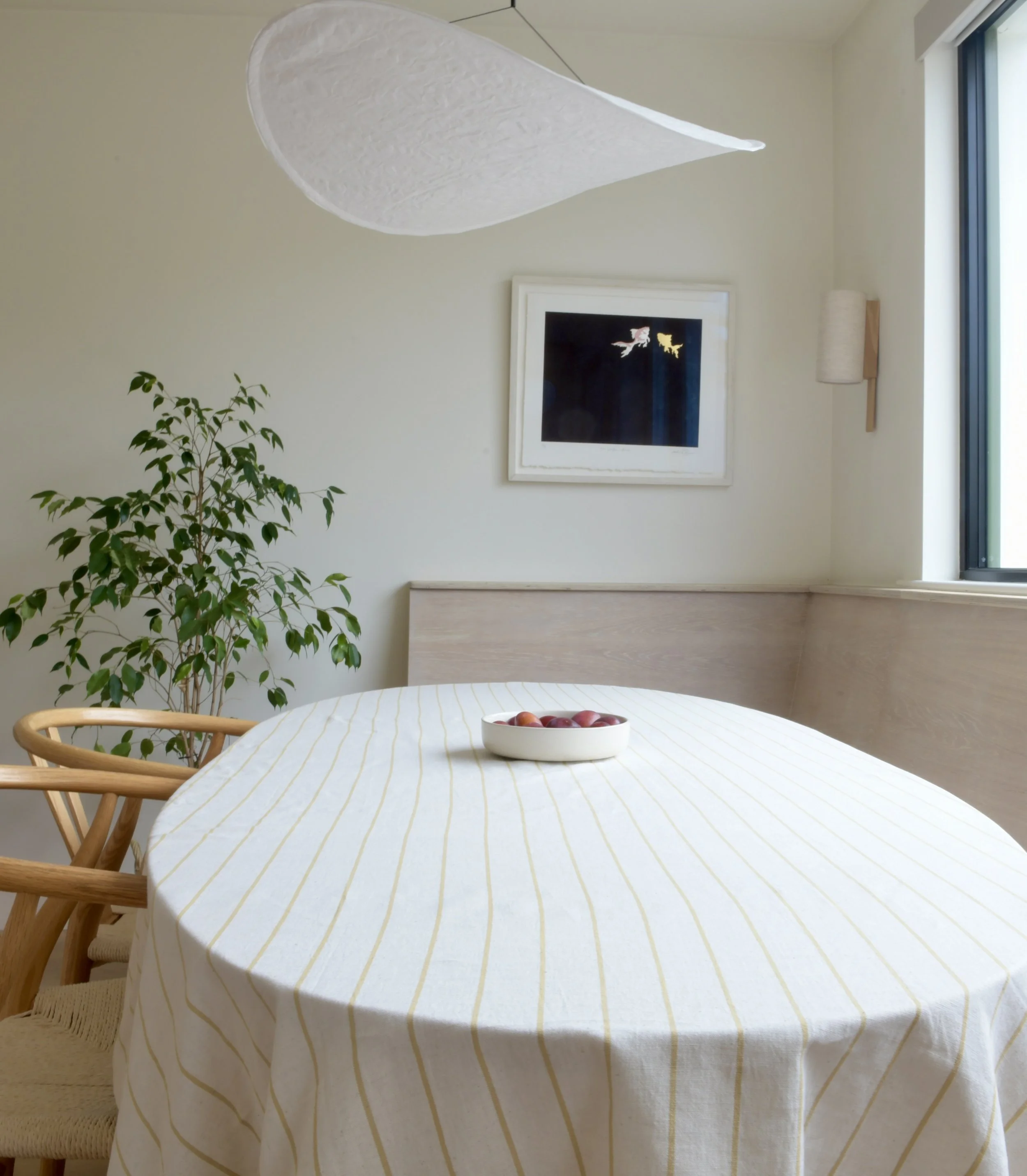 Interior view of a room with a wooden dining table covered with a yellow and white striped tablecloth, holding a white bowl with red fruit, and wooden chairs. The room has a large window with a black frame, a framed picture of two abstract animals on