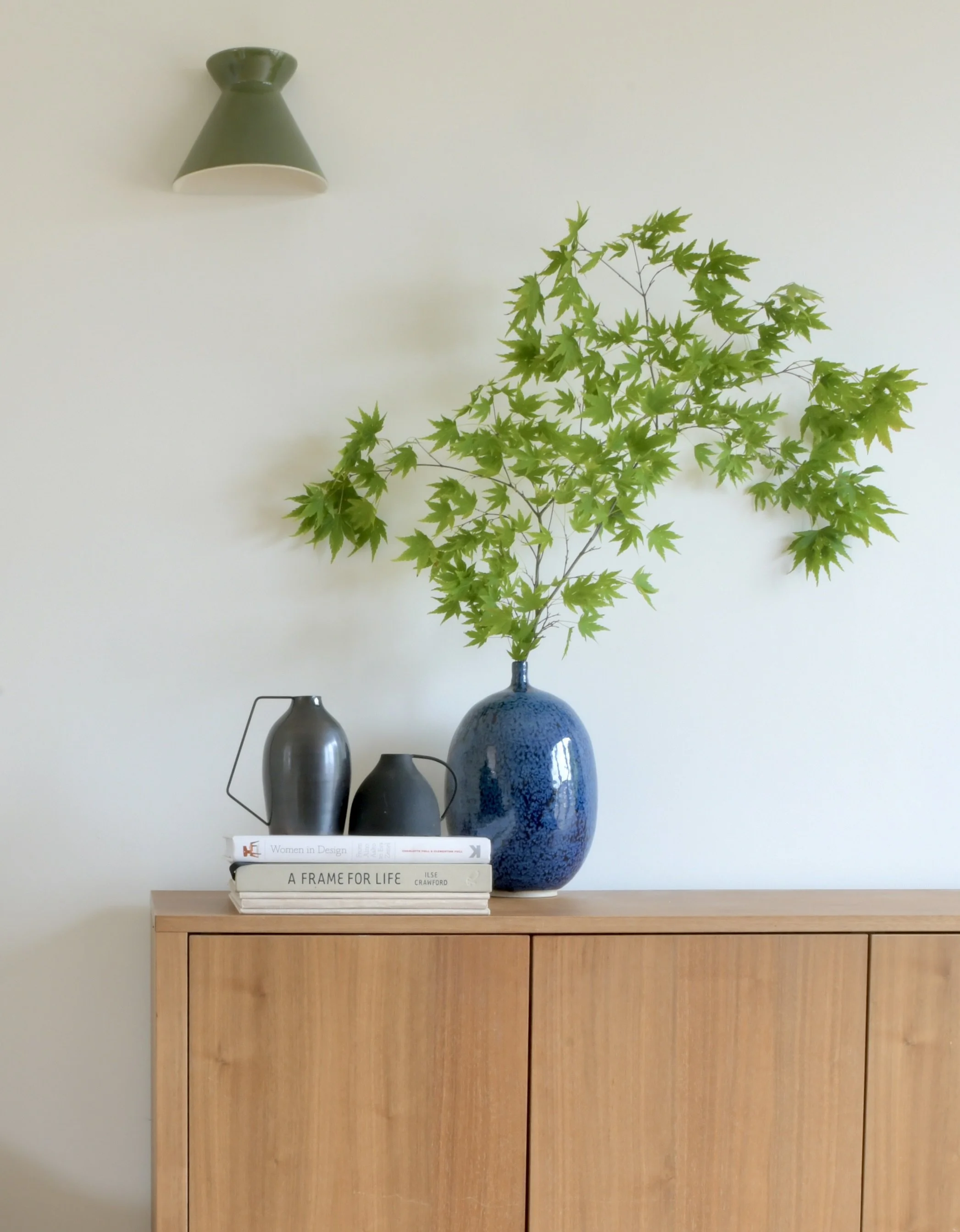 A wooden cabinet with a large blue vase holding green leafy branches on top, along with three stacked books and two small decorative vases in different shades of gray and black.