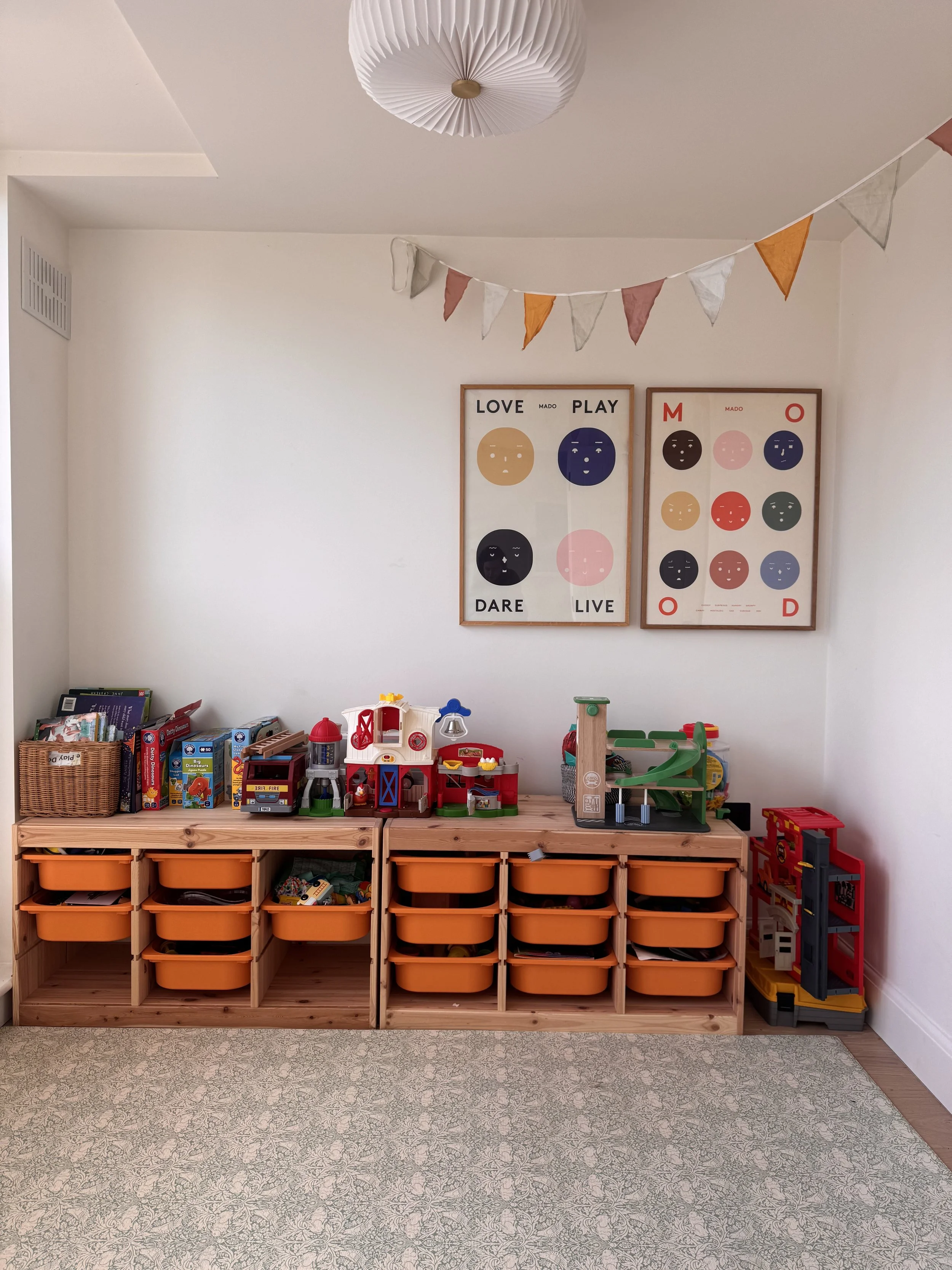 A wooden toy storage unit with multiple orange bins, filled with children's toys including toy buildings and vehicles. Above are two framed art prints with colorful faces and motivational words, and a fabric bunting banner hanging from the ceiling. T