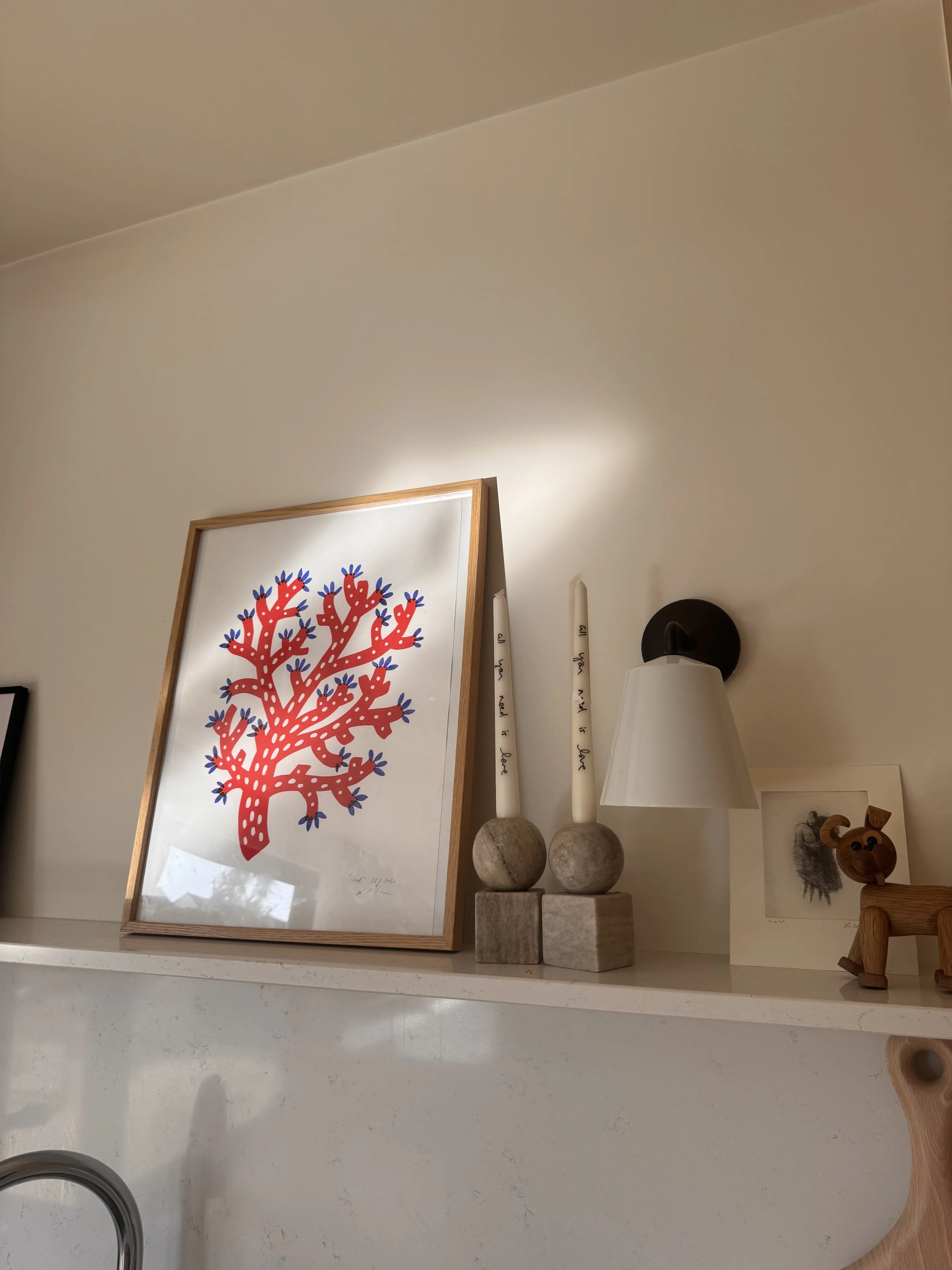 Decorative corner with framed artwork of a red and blue stylized tree, two white candles on stone and wooden bases, a white table lamp, and small wooden animal figure on wall.