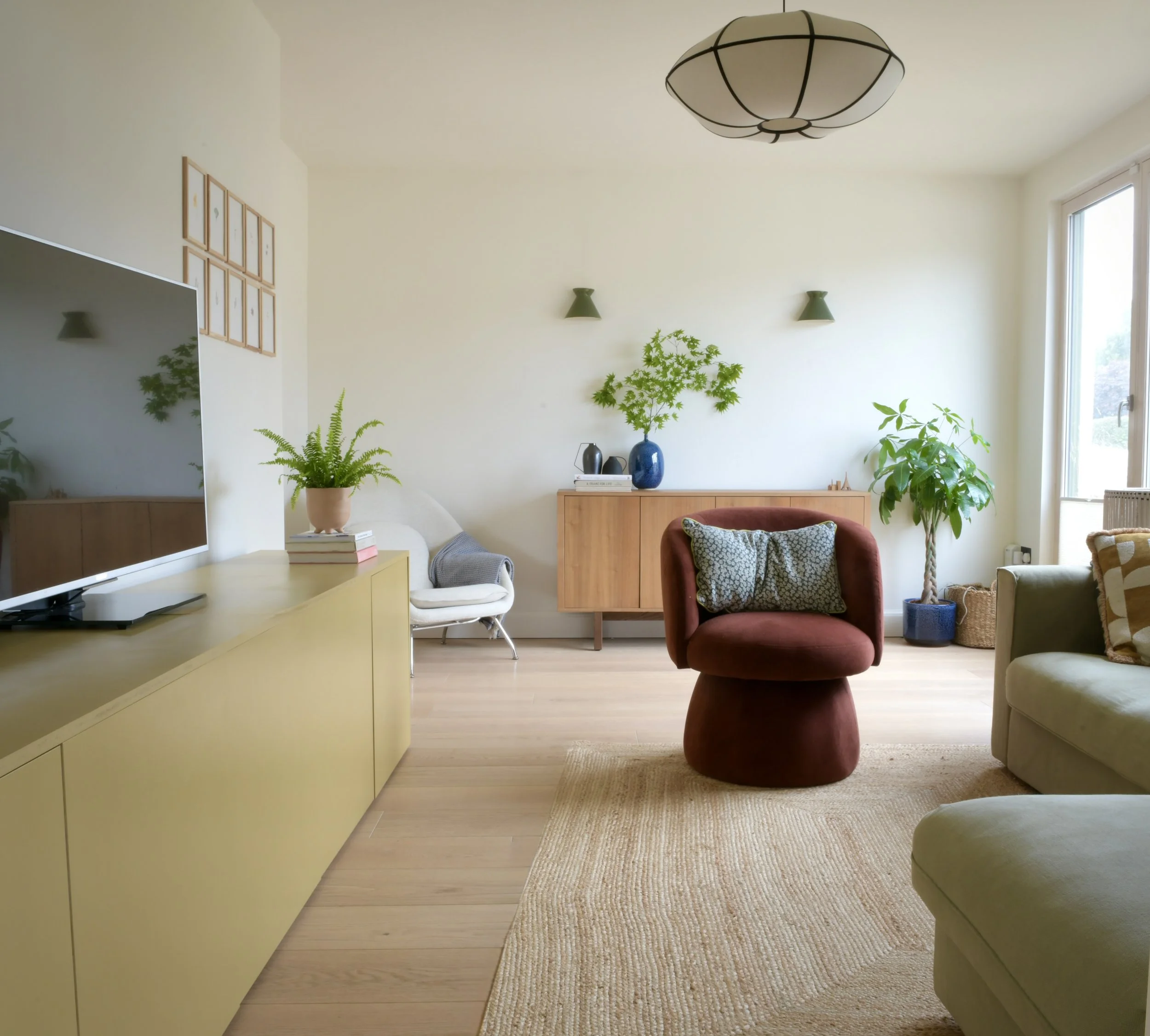 Living room with beige sofa, red velvet armchair, wooden sideboard, green plants, large window, wall-mounted lights, and ceiling lamps.