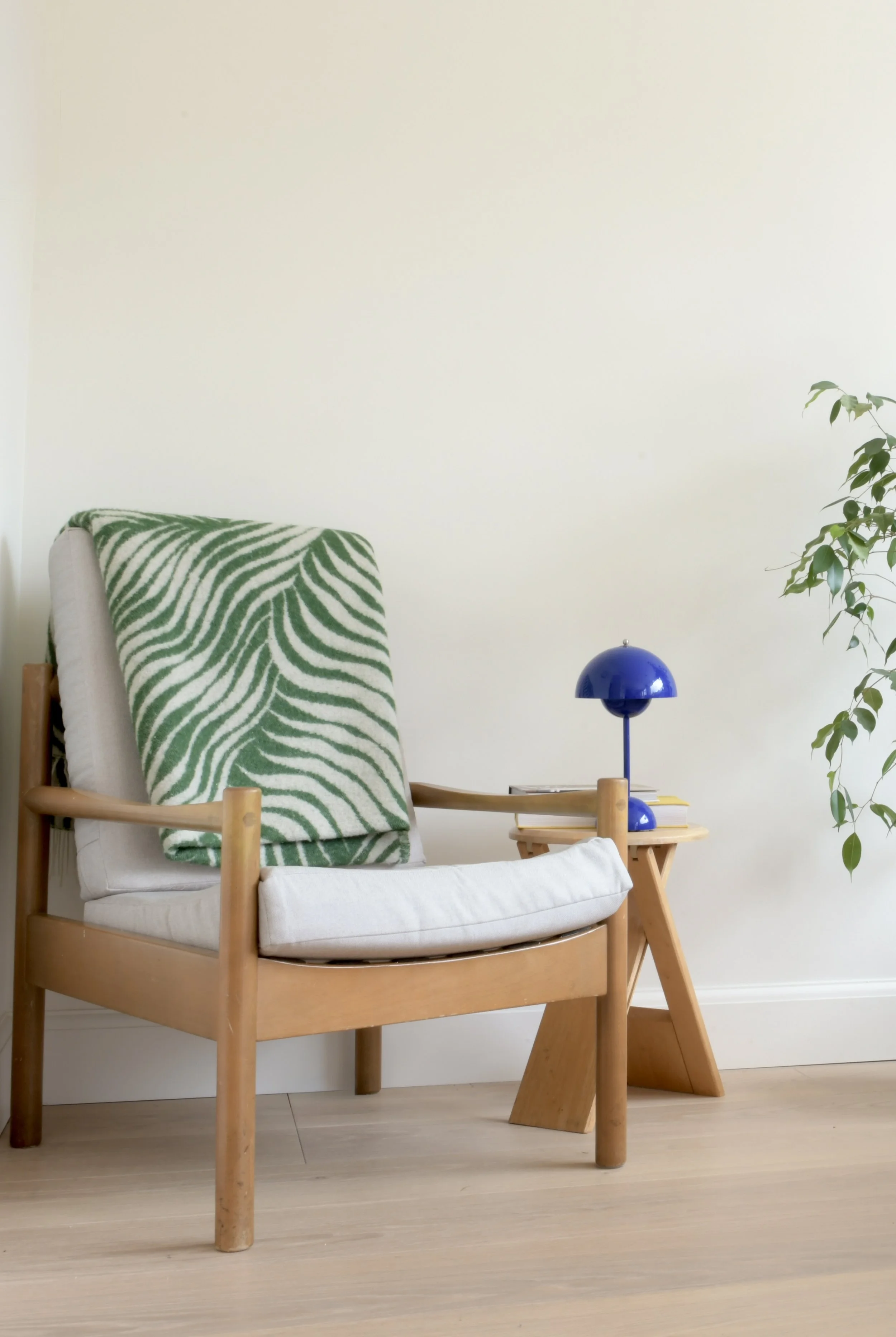 A cozy living space corner with a wooden armchair, a white cushion, a green and white leaf-patterned blanket, a small wooden side table, a blue dome-shaped table lamp, and a green leafy plant in the background.