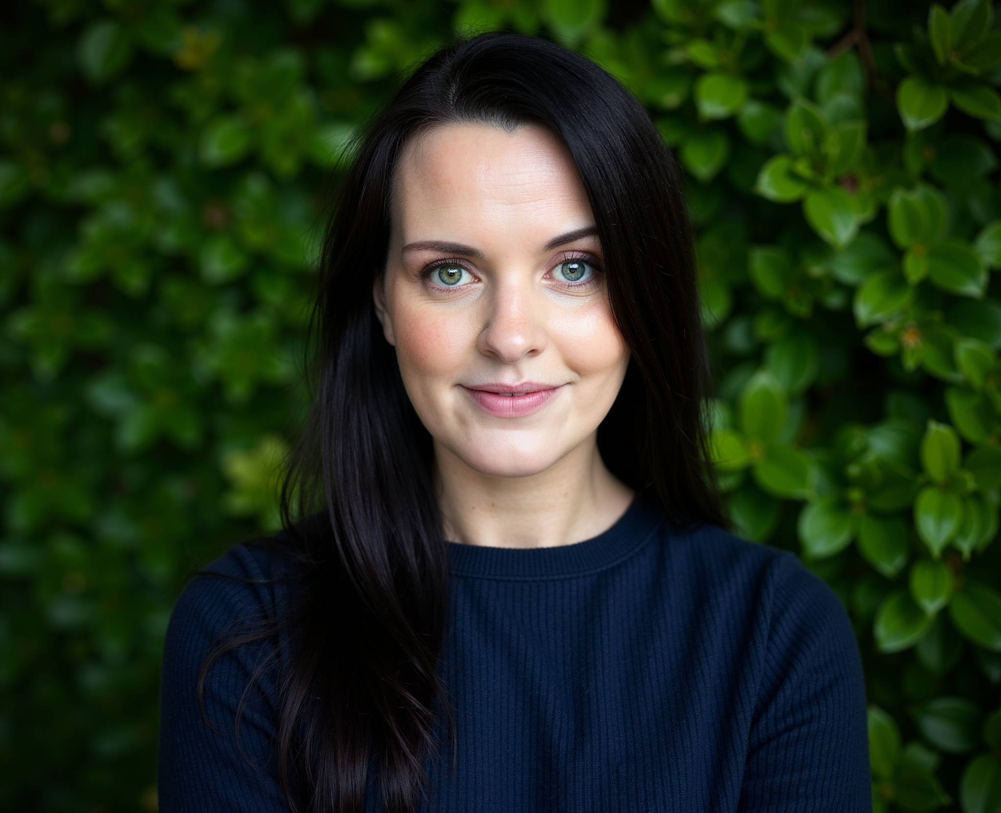A woman with long dark hair and blue eyes smiling in front of a green leafy background.