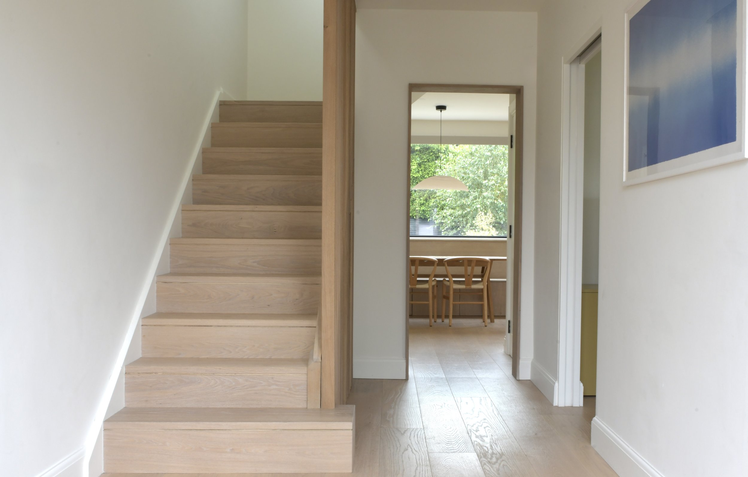 A view of an interior home space with a wooden staircase on the left, white walls, and a doorway leading to a dining area with a window showing green trees outside. There is a hanging light over the dining table.