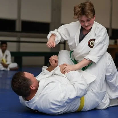 Two young boys practicing judo on a mat, one is on the ground with a yellow belt, the other is standing and wearing a green belt, engaging in a friendly match.