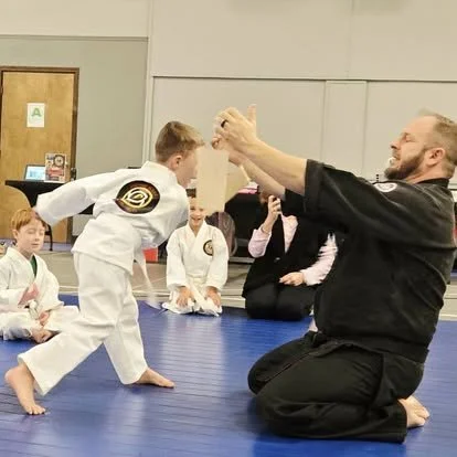 A martial arts instructor demonstrates a technique with a young student in a martial arts dojo, others in gi uniforms observe.