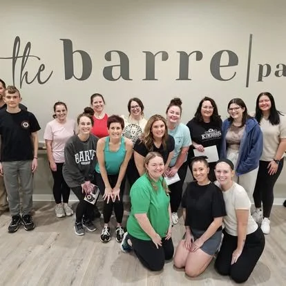 A group of 13 women and one young man posing together in front of a wall that says 'the barrel part'.