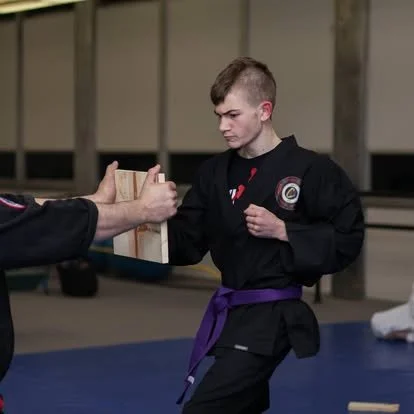 Martial arts practitioner in a black gi with a purple belt in a dojo, receiving a certificate from an instructor.