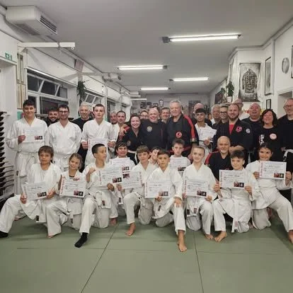 Group of children and adults in a martial arts dojo, holding certificates, dressed in white gi uniforms and black belts, posing for a photo after a training session or graduation.