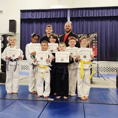Group of children in martial arts uniforms holding certificates, with an instructor in front, on a karate dojo mat.