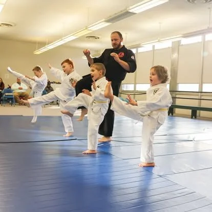 Martial arts instructor teaching children in a martial arts class, all wearing white uniforms, performing a high kick in a gym with blue mats.