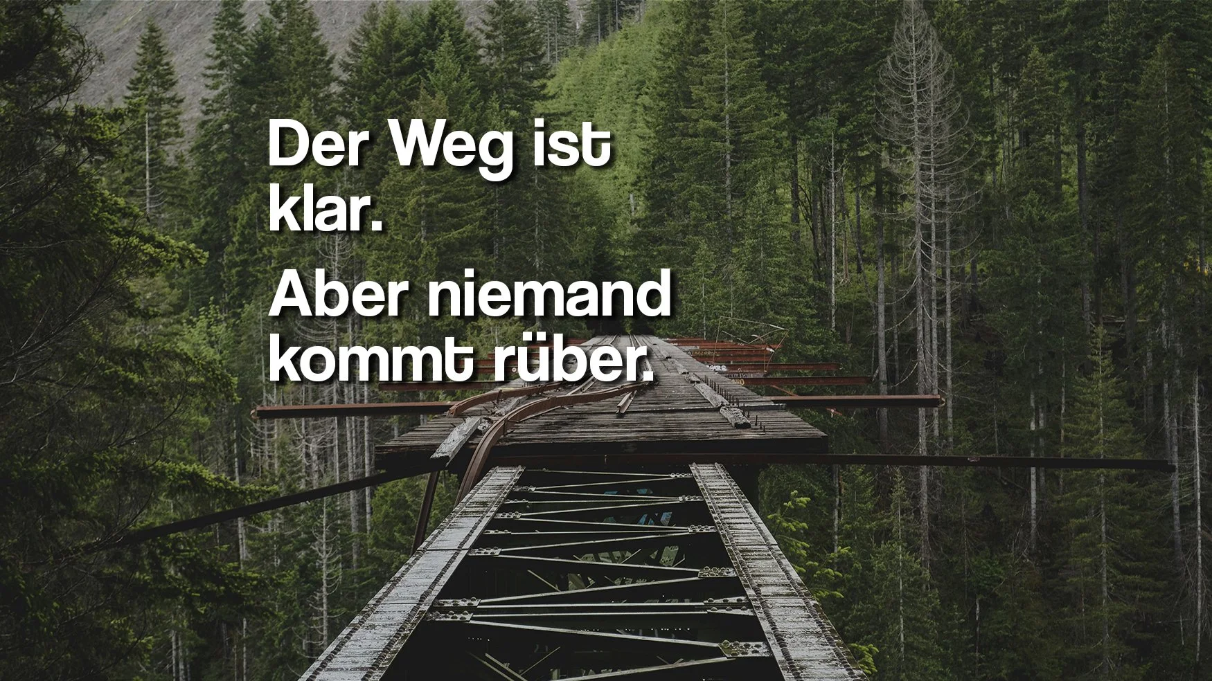 Verfallene alte Eisenbahnbrücke in einem Wald mit grünen Bäumen und einem Berg im Hintergrund, mit überlagertem deutschen Text: 'Der Weg ist klar. Aber niemand kommt rüber.'