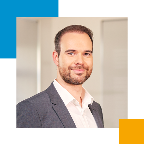 Ein Mann trägt einen grauen Anzug und ein weißes Hemd, steht in einem Büroumfeld.