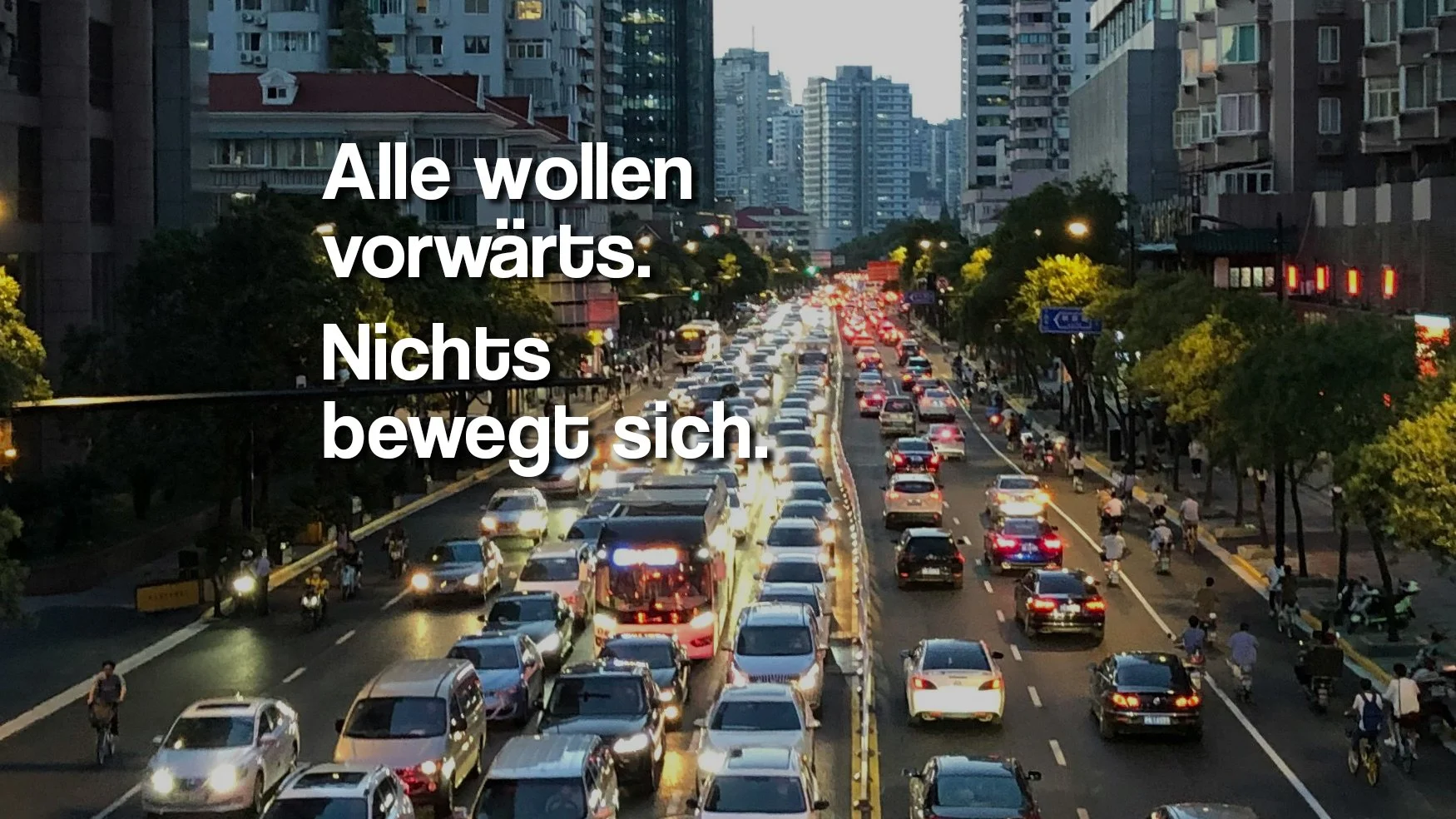 Stadtverkehr mit Stau auf einer mehrspurigen Straße, hohe Gebäude im Hintergrund, Abendzeit, Beleuchtung auf den Straßen und in den Gebäuden.